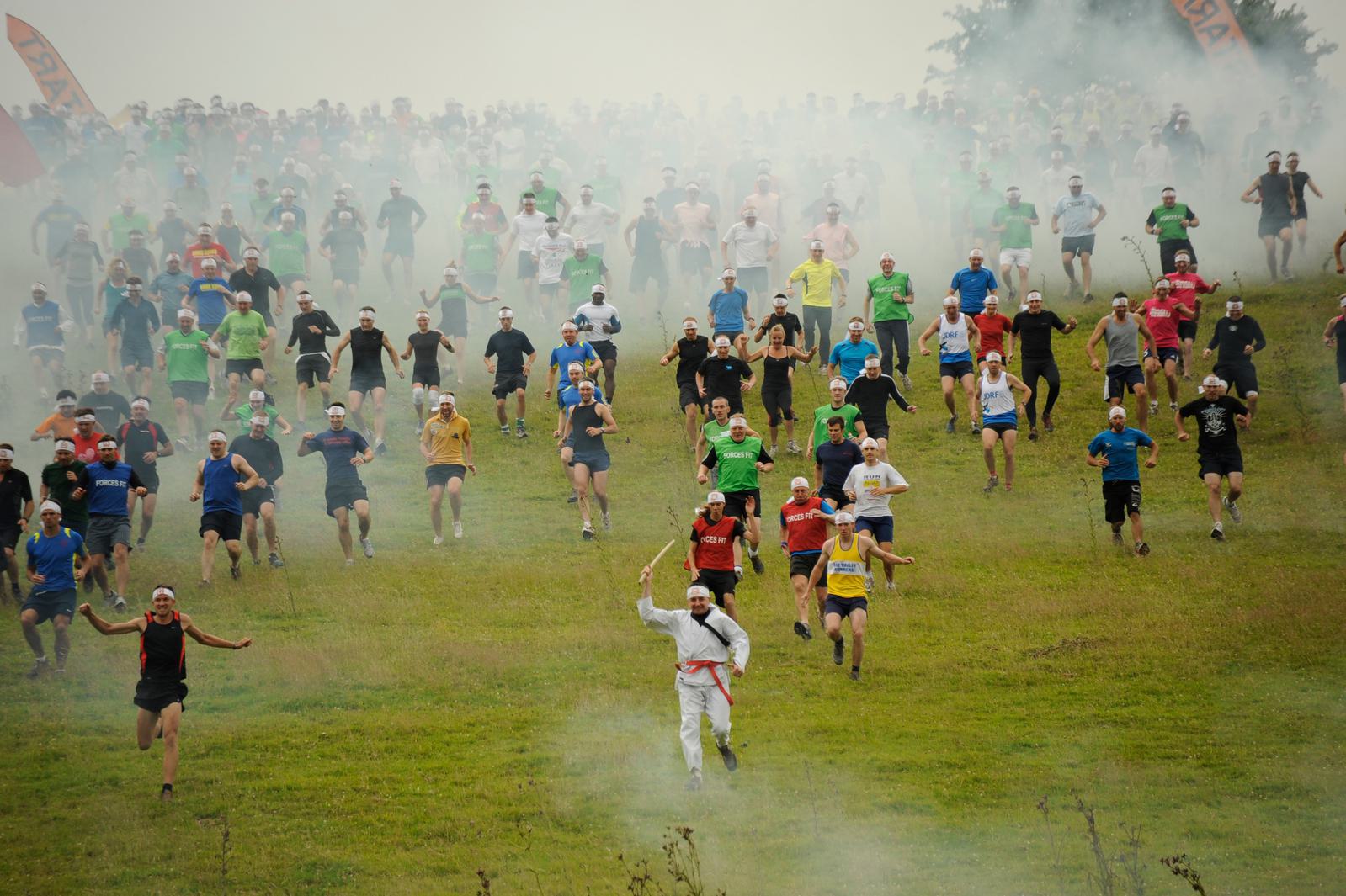 A large group of people in colorful athletic wear race downhill on a grassy field, with some smoke in the background. A person dressed in white and red leads at the front, holding an object in the air. It appears to be an energetic, competitive event.