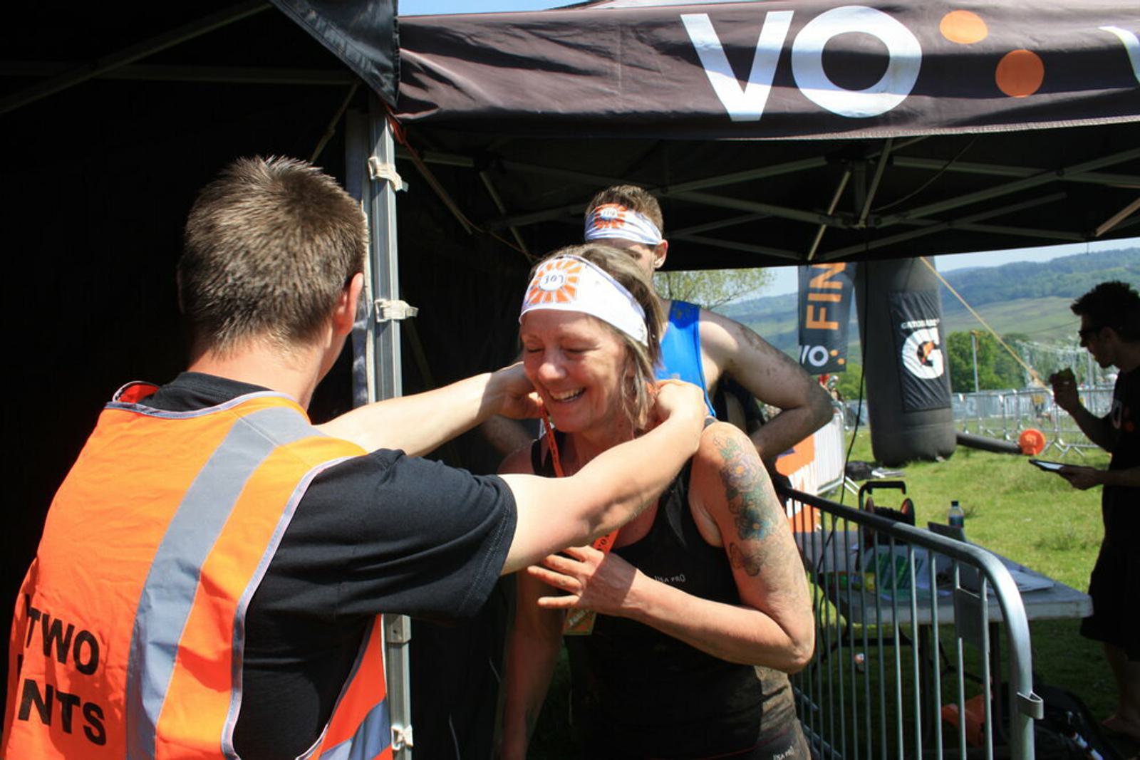 A woman wearing a headband is receiving a medal from a man in a high-visibility vest at the finish line of an outdoor event. She appears joyful and emotional. Other participants and part of the event setup are visible in the background.