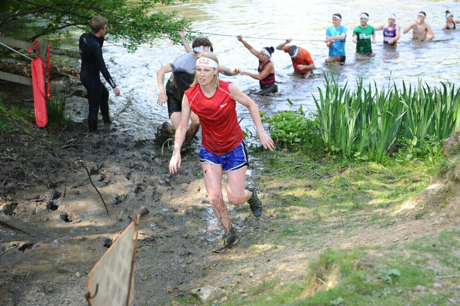 A woman in a red top and blue shorts, covered in mud, runs out of the water during an outdoor obstacle race. Behind her, several other participants wade through the water near the shore, with some still in the water and others just exiting.