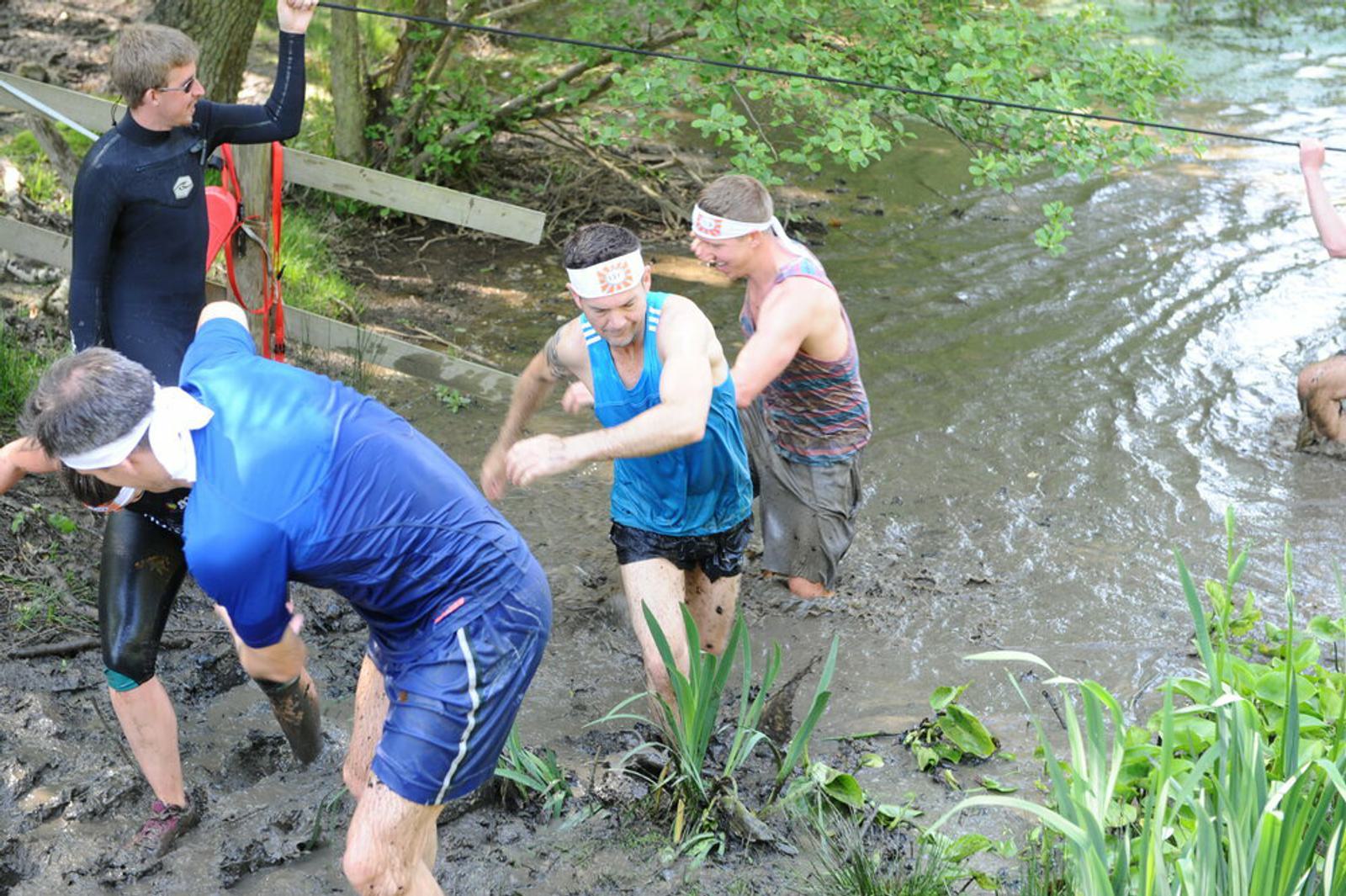 Four individuals are wading through a muddy water obstacle course in a wooded area. They are dressed in athletic gear and wearing headbands. Some are using ropes for support, while another is helping a teammate out of the mud. The scene looks challenging but fun.