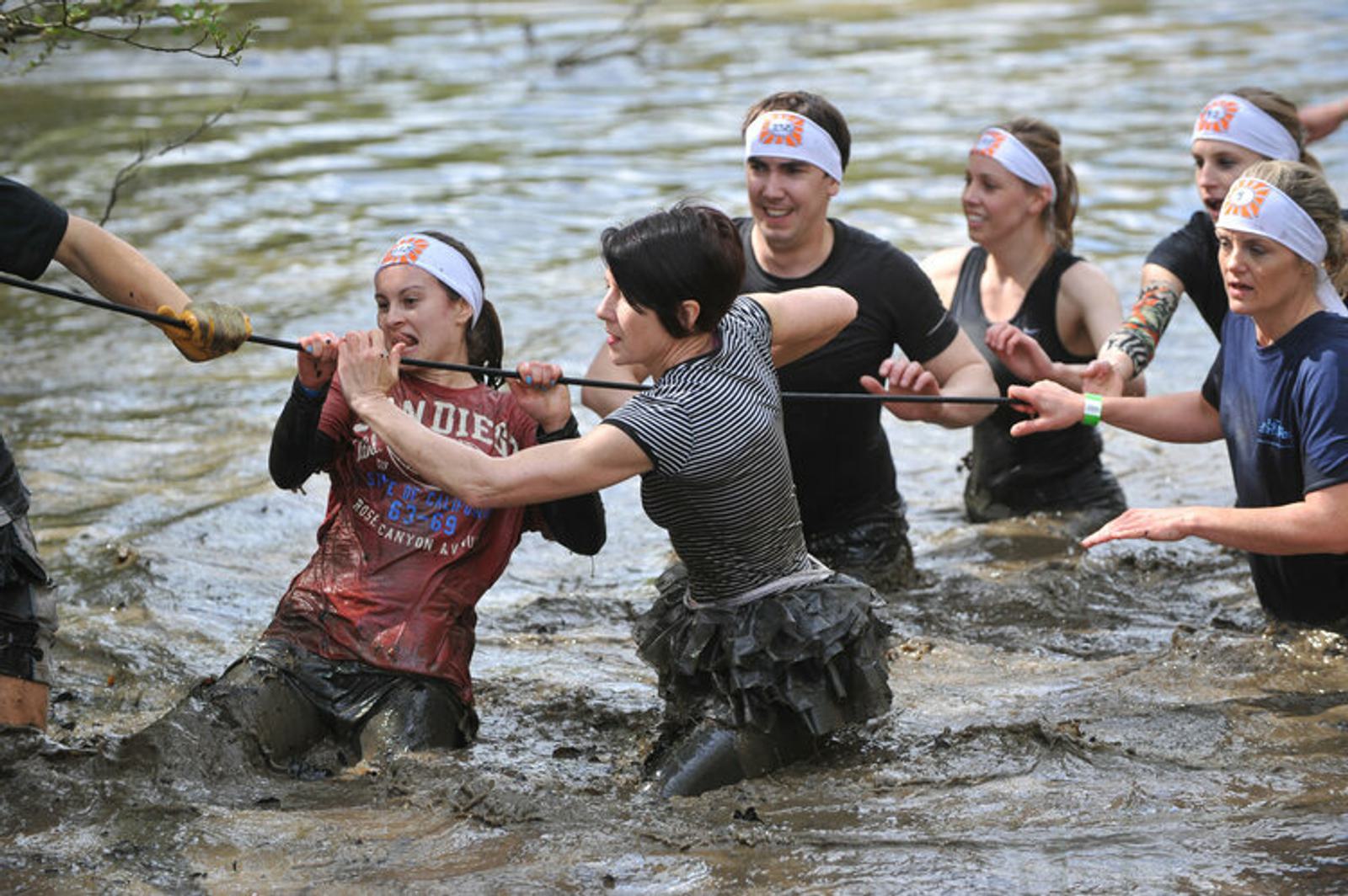 A group of people participating in a mud run is seen wading through a muddy watercourse. One woman in a red shirt is being assisted by others using a rope. They are wearing headbands and appear to be having fun despite the challenging conditions.