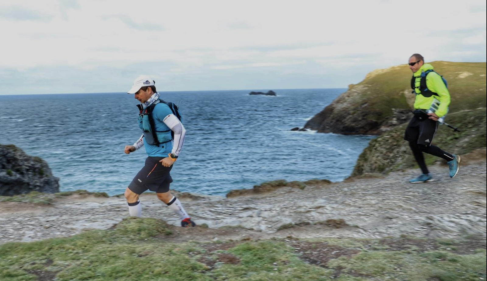 Two runners jog along a rocky coastal path with the ocean and cliffs in the background. The runner in the foreground wears a white cap and blue shirt, while the runner behind wears a bright green jacket. Both carry hydration packs and run in steady stride.