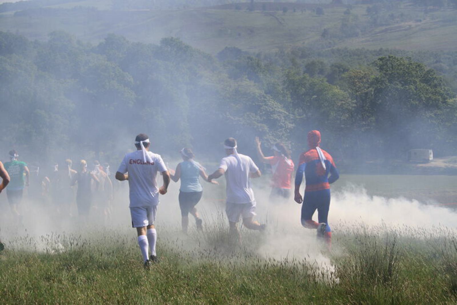 A group of people, including one dressed in a superhero costume, run through a grassy field with patches of white smoke or mist. Trees and a hilly landscape are visible in the background, and a sunny sky illuminates the scene.