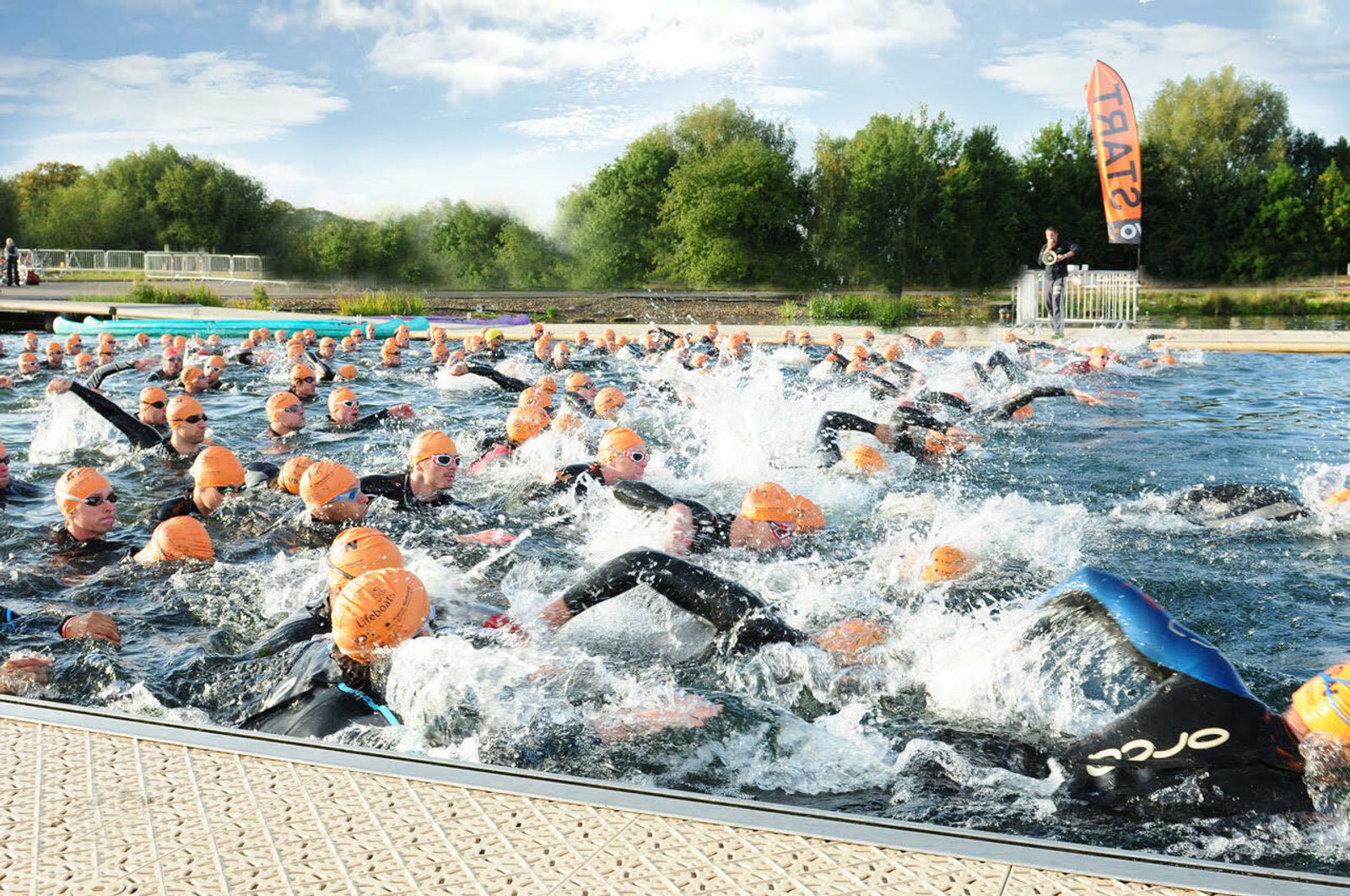 A large group of swimmers wearing black wetsuits and orange swim caps start a race in open water. The foreground shows churning water, and the background features lush green trees under a partly cloudy sky. A "Start" banner is visible on the right side.