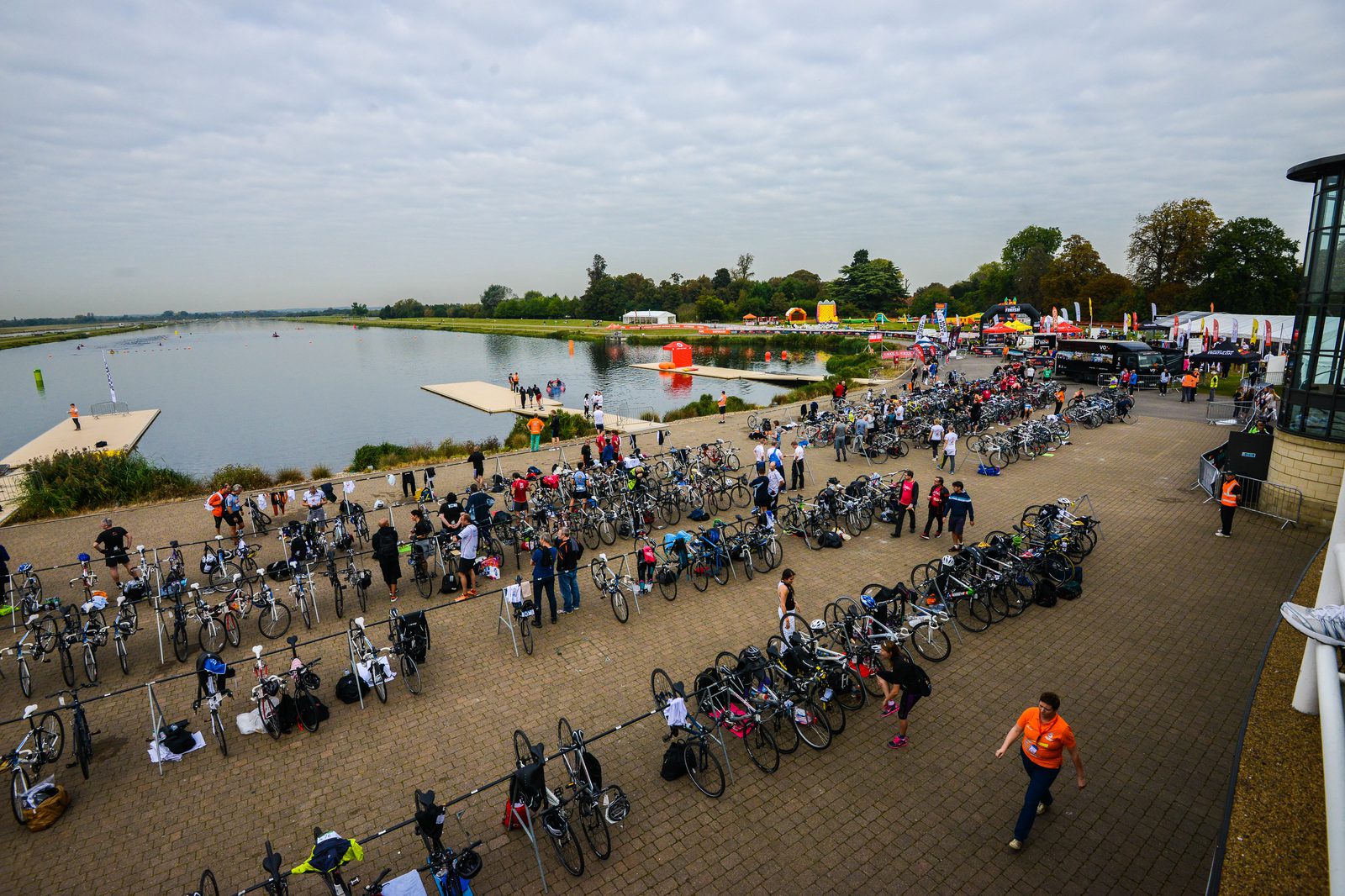 An overhead view of a triathlon transition area beside a body of water. Numerous bicycles are parked in rows, and participants are preparing for the next stage of the race, with some walking and others adjusting their gear. The sky is overcast.
