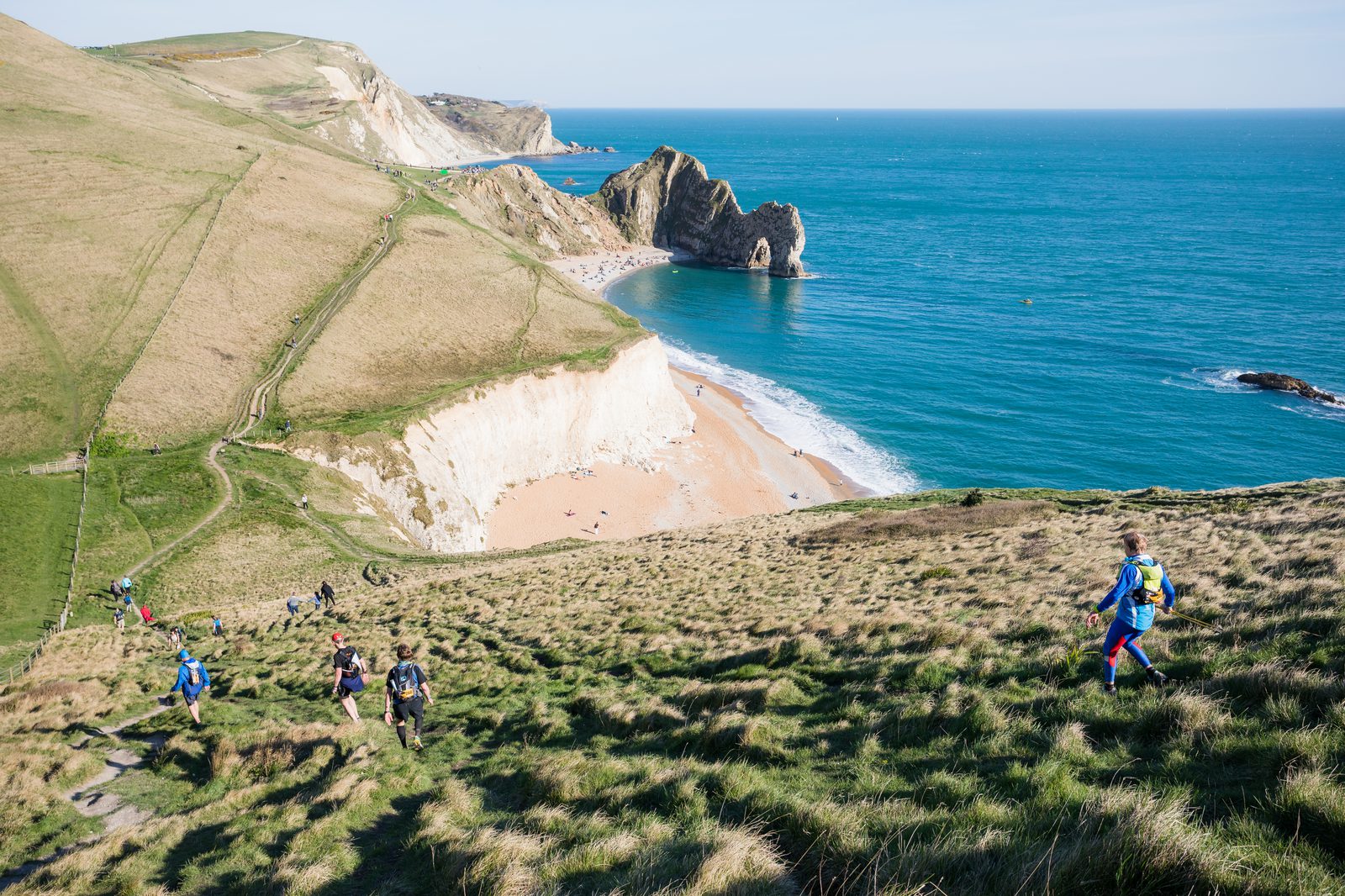 Hikers walk along grassy hills overlooking a coastline with cliffs and a sandy beach. The clear blue sea contrasts with the greenery of the land, and an arch-shaped rock formation is visible in the distance. The sky is clear, indicating a sunny day.