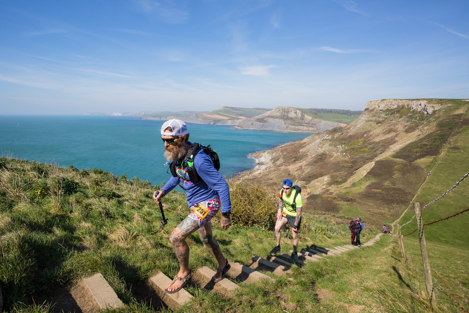 Trail runners ascend a steep set of steps along a coastal path overlooking the ocean and cliffs. The lead runner wears a blue shirt, cap, and backpack, while the runners behind wear bright clothing. The sky is clear and the scenery is vibrant with green and brown hues.