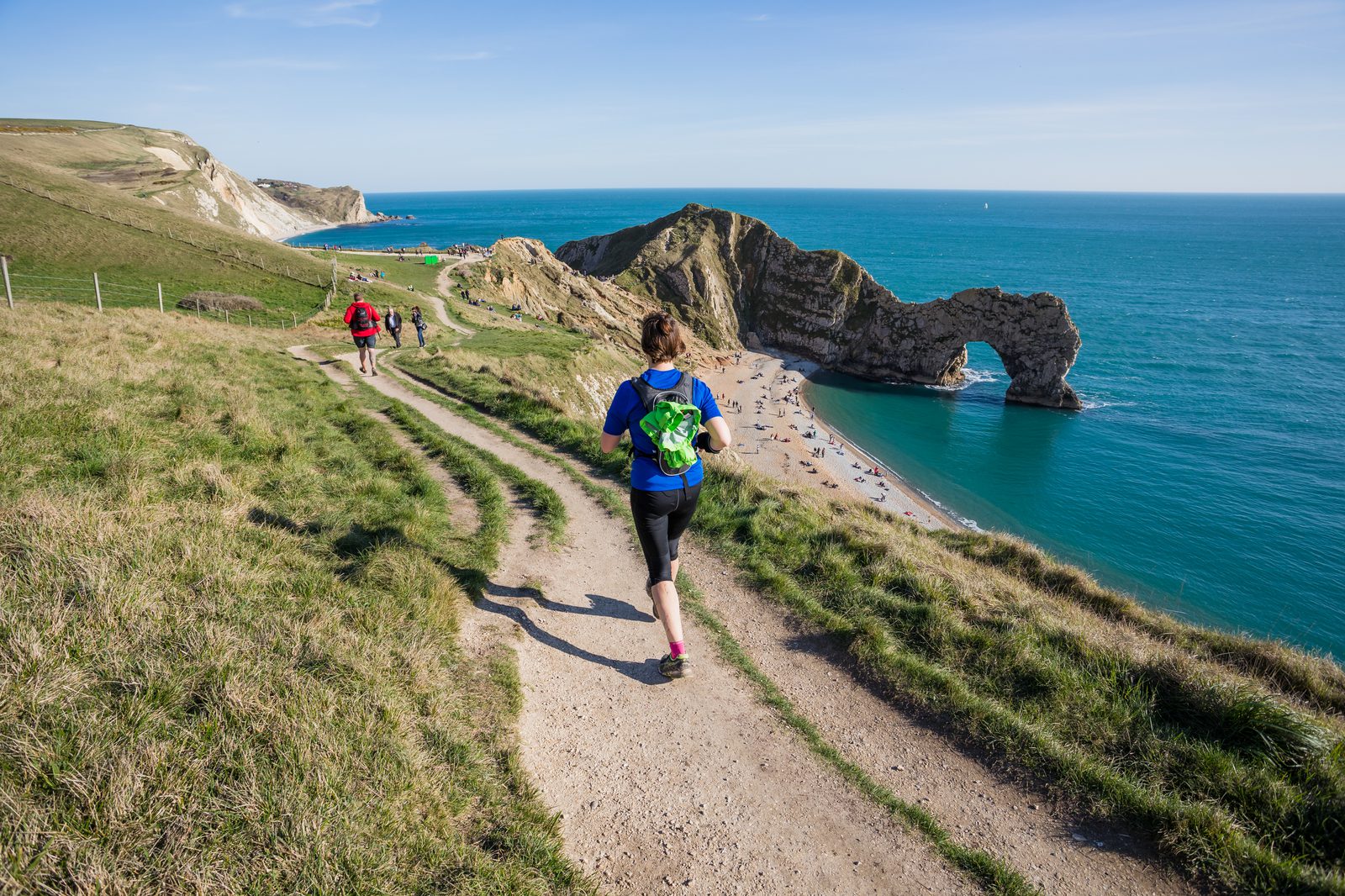A group of people are trail running on a grassy path towards the iconic Durdle Door arch on the Jurassic Coast in Dorset, England. The clear blue sky and turquoise sea provide a picturesque backdrop, with sandy beach visible below the arch.