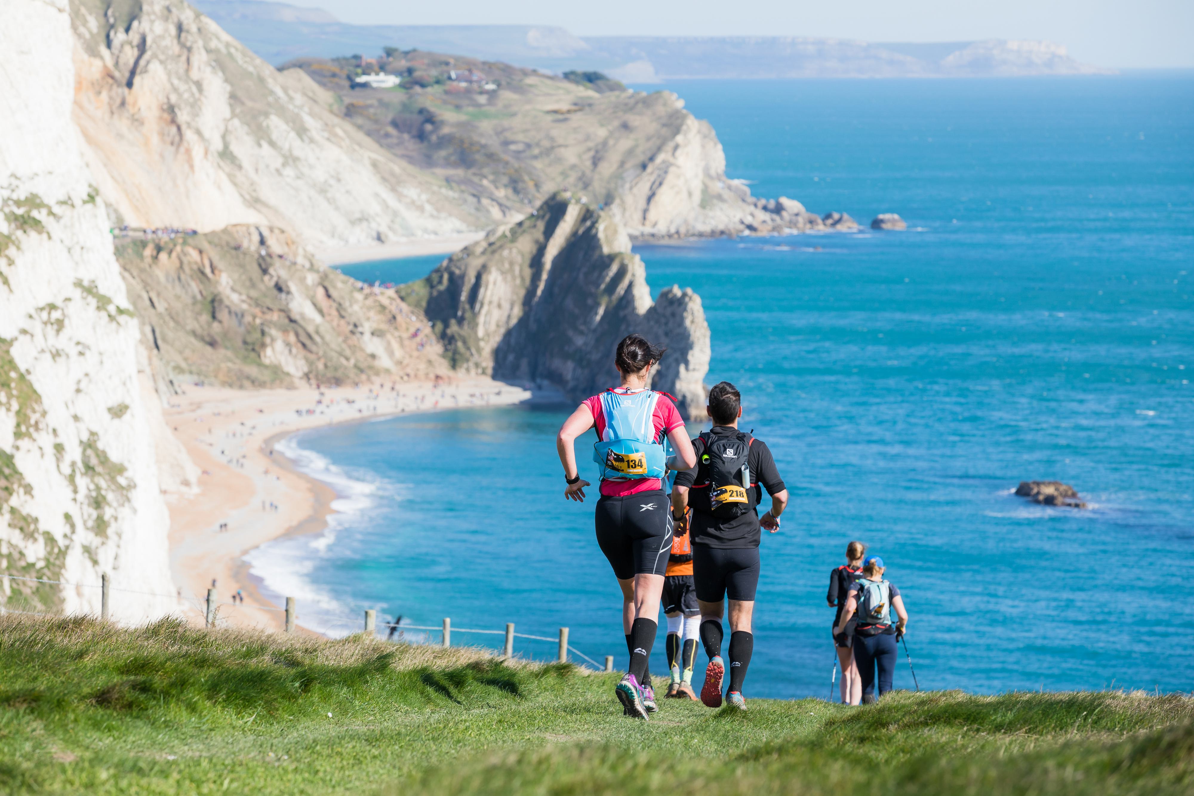 Three people are trail running on a grassy path along a coastal cliff with a scenic view of the ocean and rugged cliffs. The sea is calm, and the sky is clear. A sandy beach and more cliffs are visible in the background.