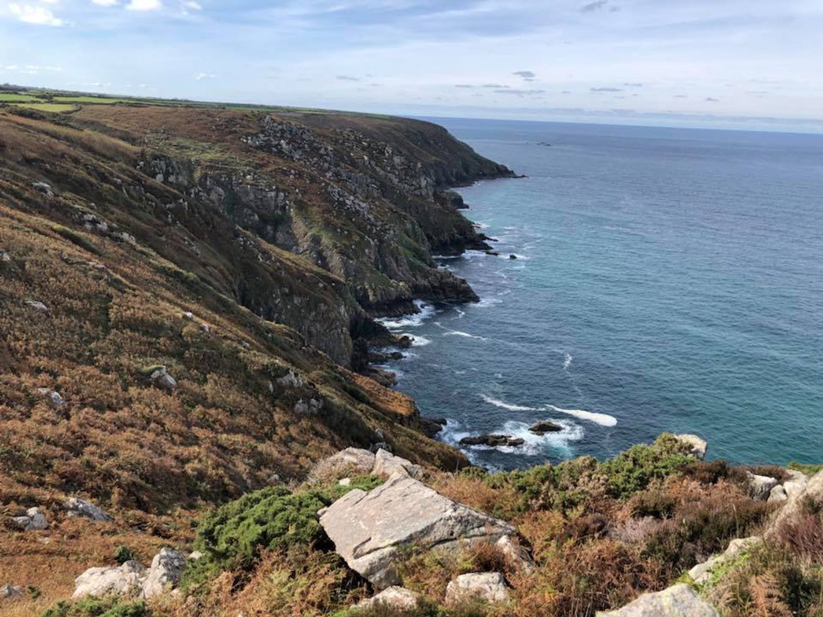 A coastal cliff lined with rocky formations and patches of grass slopes down to the turquoise ocean waters below. The waves gently lap against the shore, and the horizon stretches out under a partly cloudy sky. The landscape is lush with green and brown vegetation.