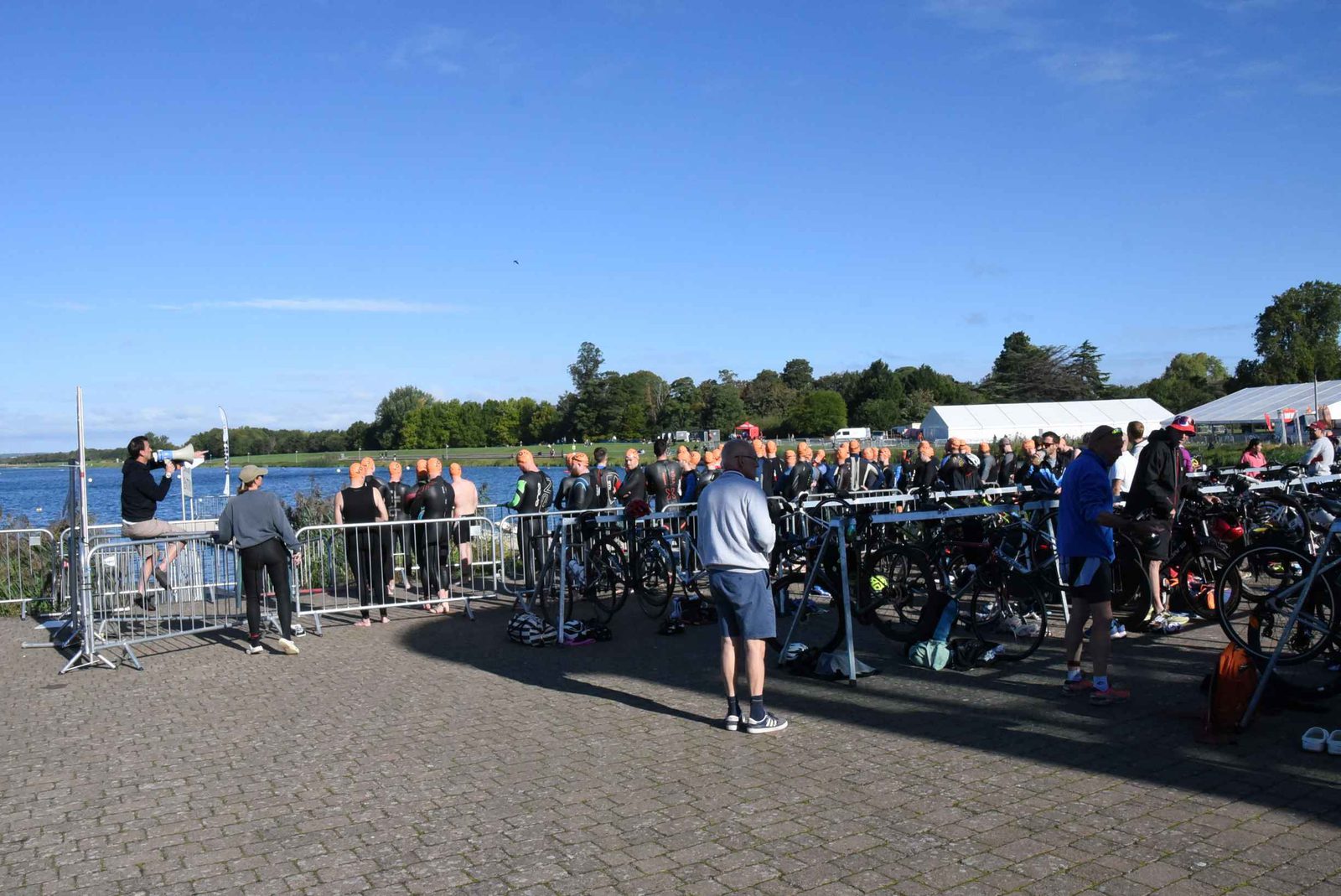 Athletes in wetsuits stand in a group near a lake, preparing for a swim; bicycles line the transition area and people watch, under a clear blue sky with trees and tents in the background.