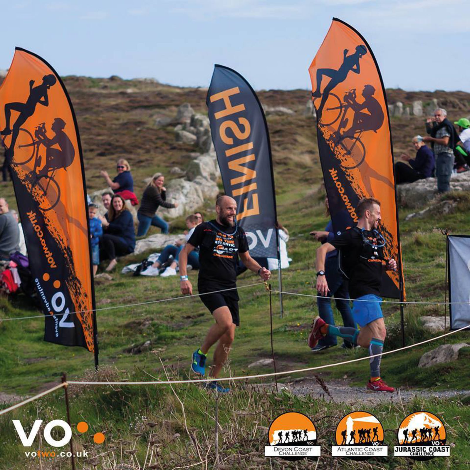 Two runners approach a race finish line marked by orange flags on a hillside trail. Spectators sit on the grass, cheering them on. The terrain appears rocky and green under a partly cloudy sky. Various race banners and logos are visible, including VO2 Multisport.