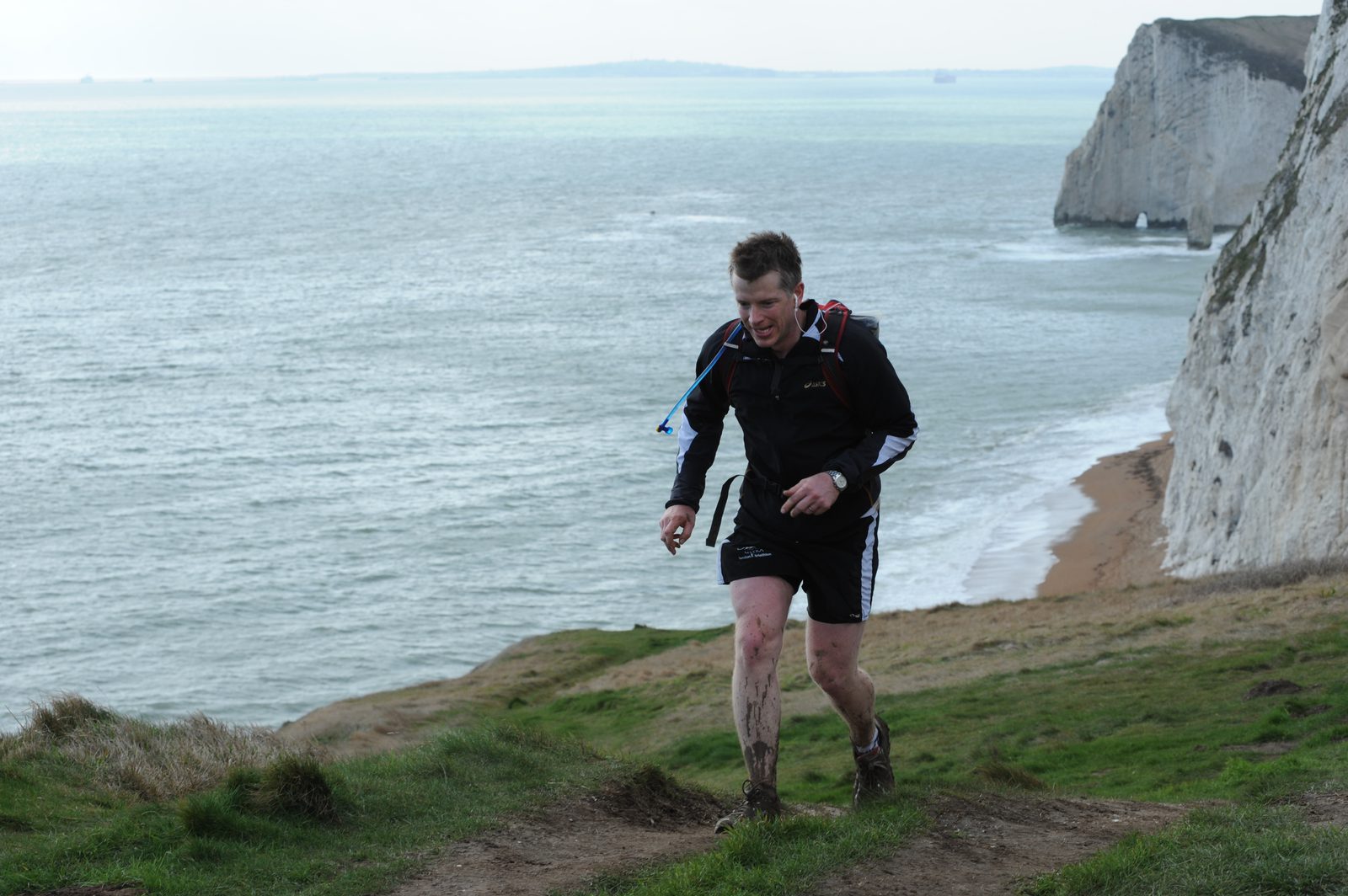A man in athletic gear runs uphill along a grassy cliffside trail beside the ocean. He has a backpack and his shoes are muddy. The background features the sea, a distant landmass, and white cliffs.