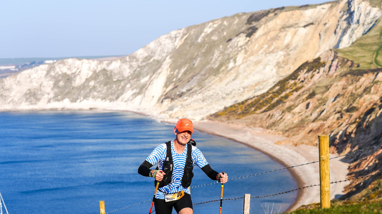 A person wearing an orange cap and striped blue shirt is hiking with trekking poles along a coastal path. The background features a steep, rocky cliff and a calm, blue sea under a clear sky.