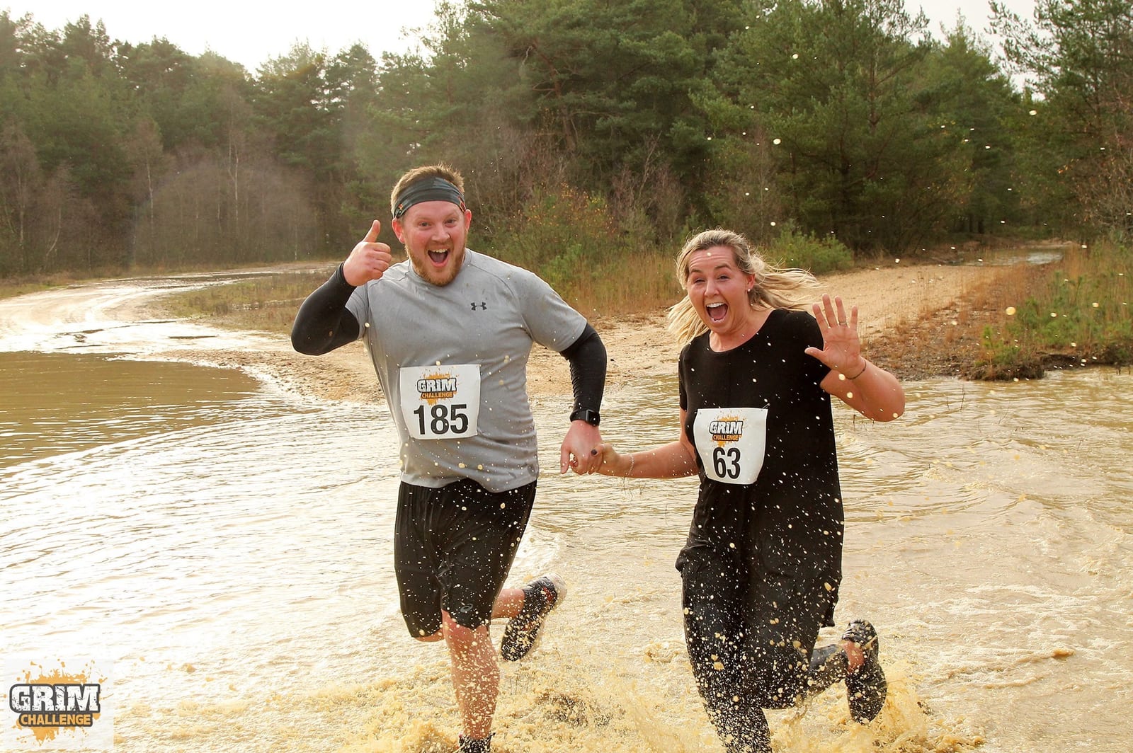 Two participants, wearing race numbers 185 and 63, run hand-in-hand through a muddy water pit in an outdoor race event. They are smiling and seem excited, with the man giving a thumbs-up and the woman waving energetically. Trees and a dirt path are visible in the background.