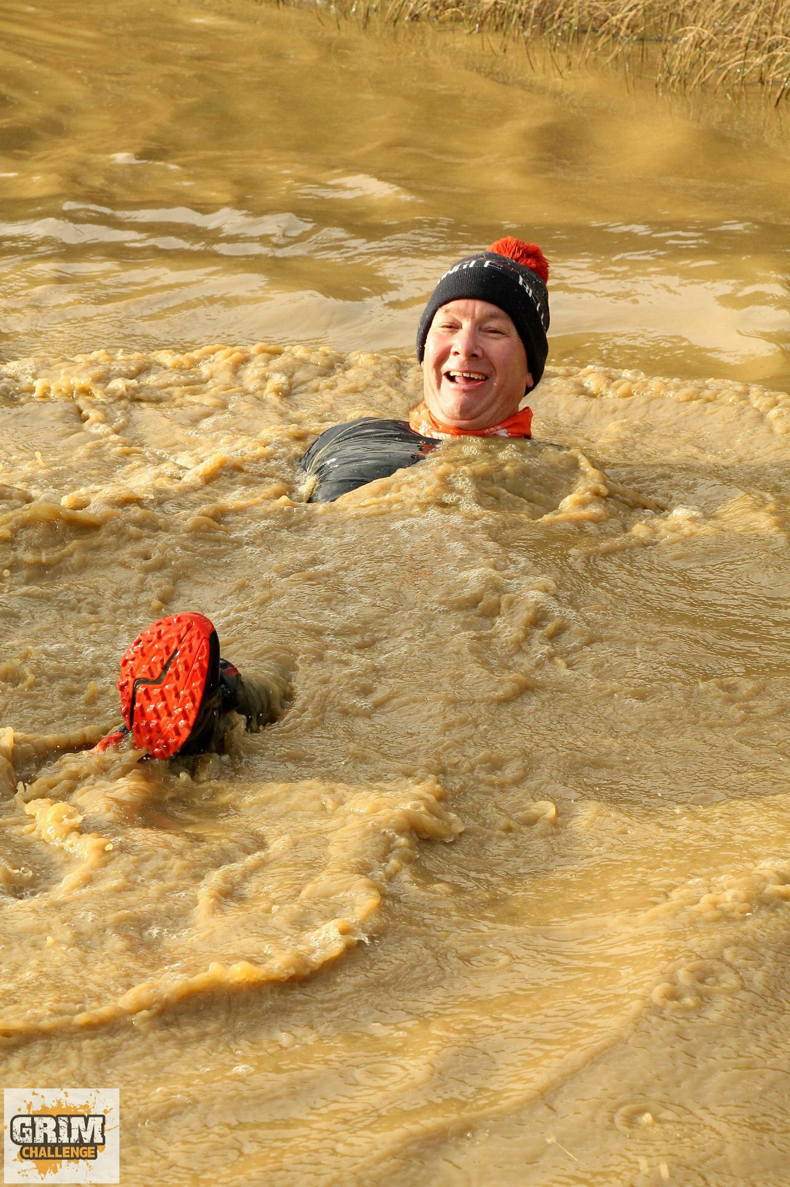 A person wearing a black beanie with a red pom-pom and black athletic gear is submerged in muddy water, smiling. Only their head and one raised muddy shoe are visible. A "Grim Challenge" logo is present in the bottom left corner.
