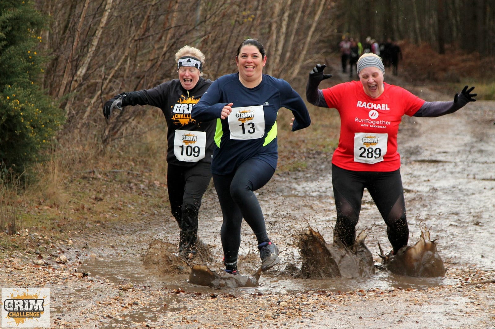 Three participants are running through a muddy trail during an outdoor race. The runner in the center wears a blue top and black pants, while the other two wear black and red tops with race bibs. All are smiling and splashing water and mud as they run. Trees line the background.