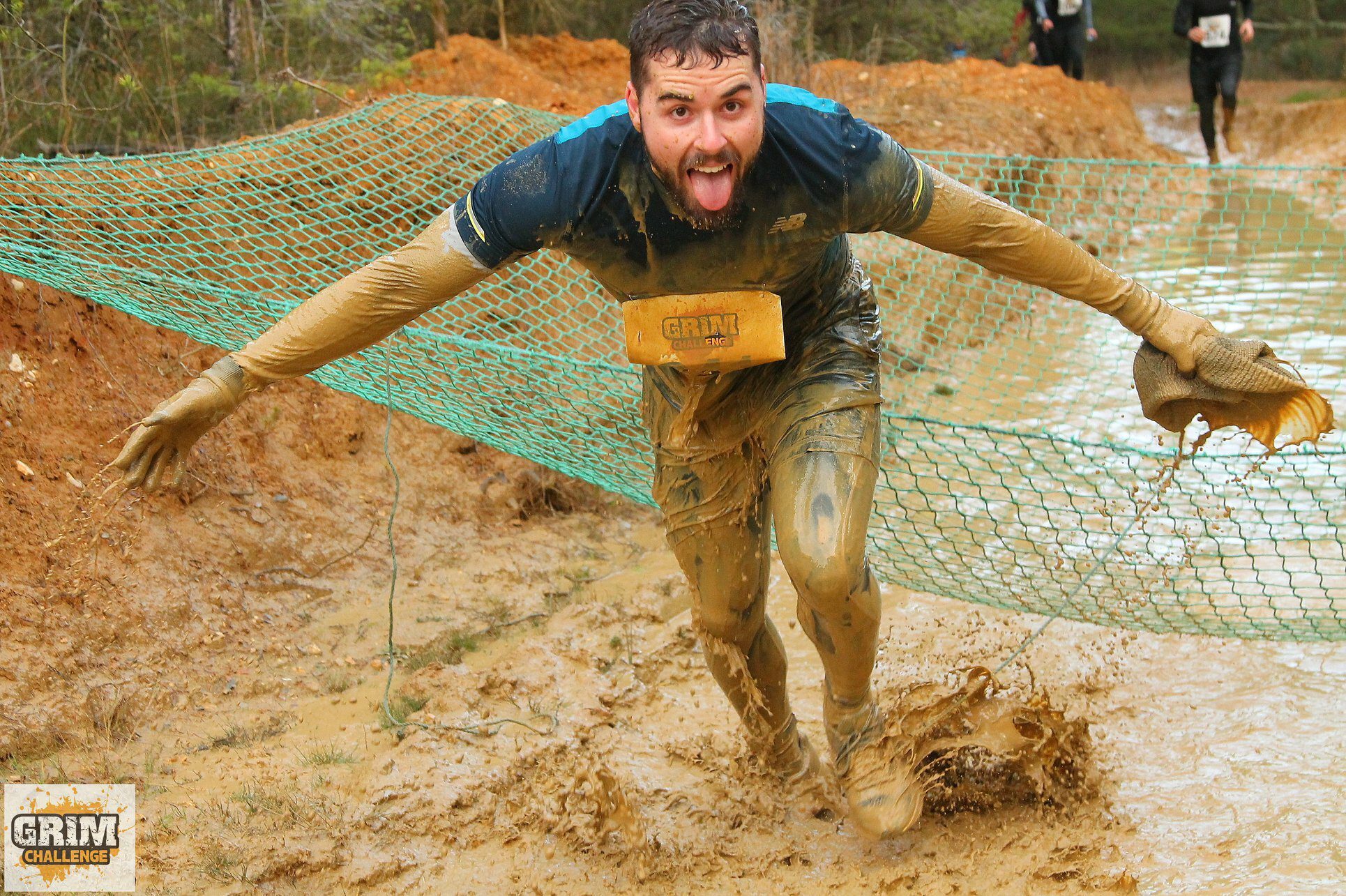 A man covered in mud is enthusiastically participating in an obstacle course race. He is crawling under a green net and has a joyful expression with his tongue out. The ground is muddy, and he is splashing through it. The race bib reads "GRIM CHALLENGE.