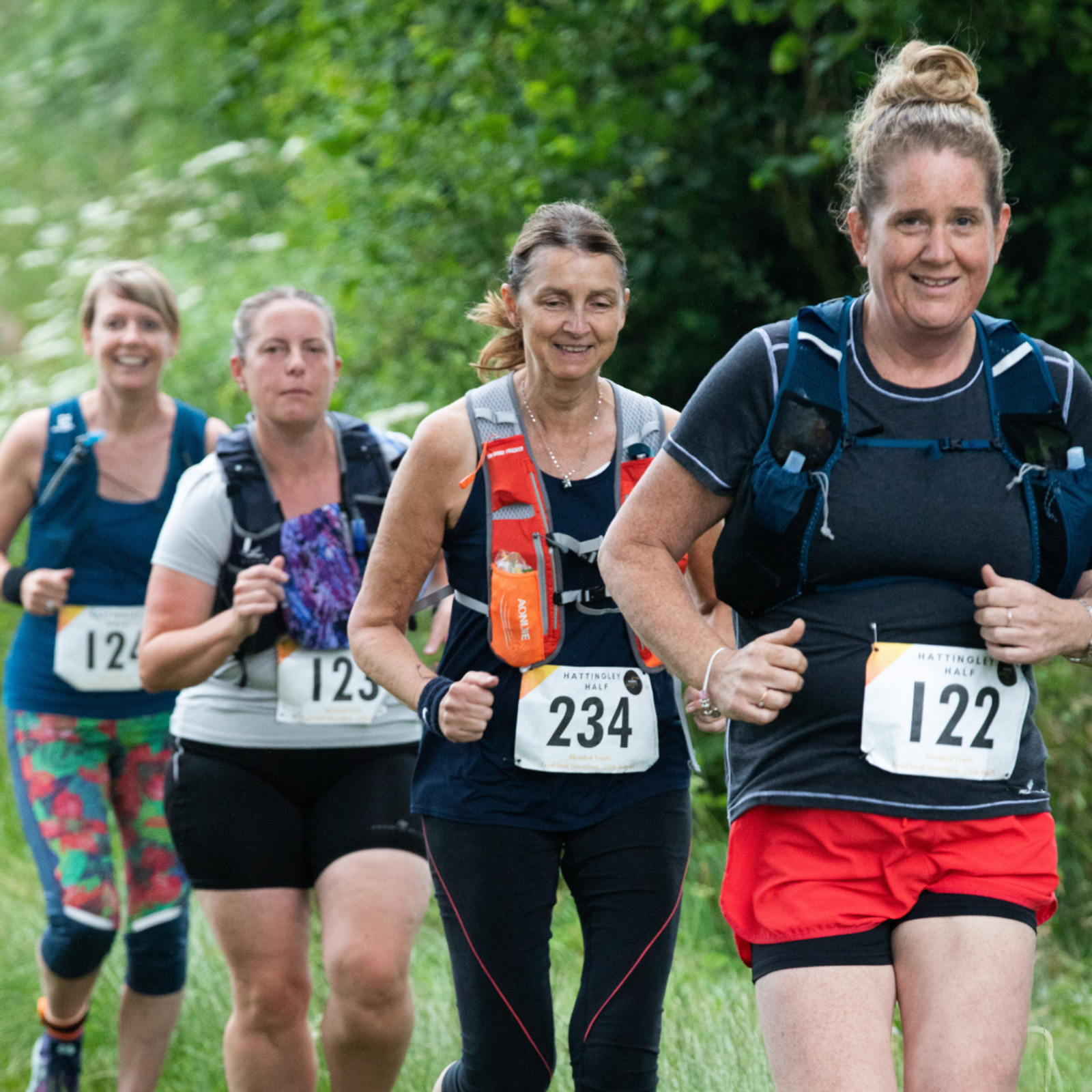 Four women are running outdoors on a trail, wearing race bibs and hydration packs. The runner in the foreground is smiling and wearing a black t-shirt with red shorts, bib number 122. The background is lush and green, hinting at a forested or park area.