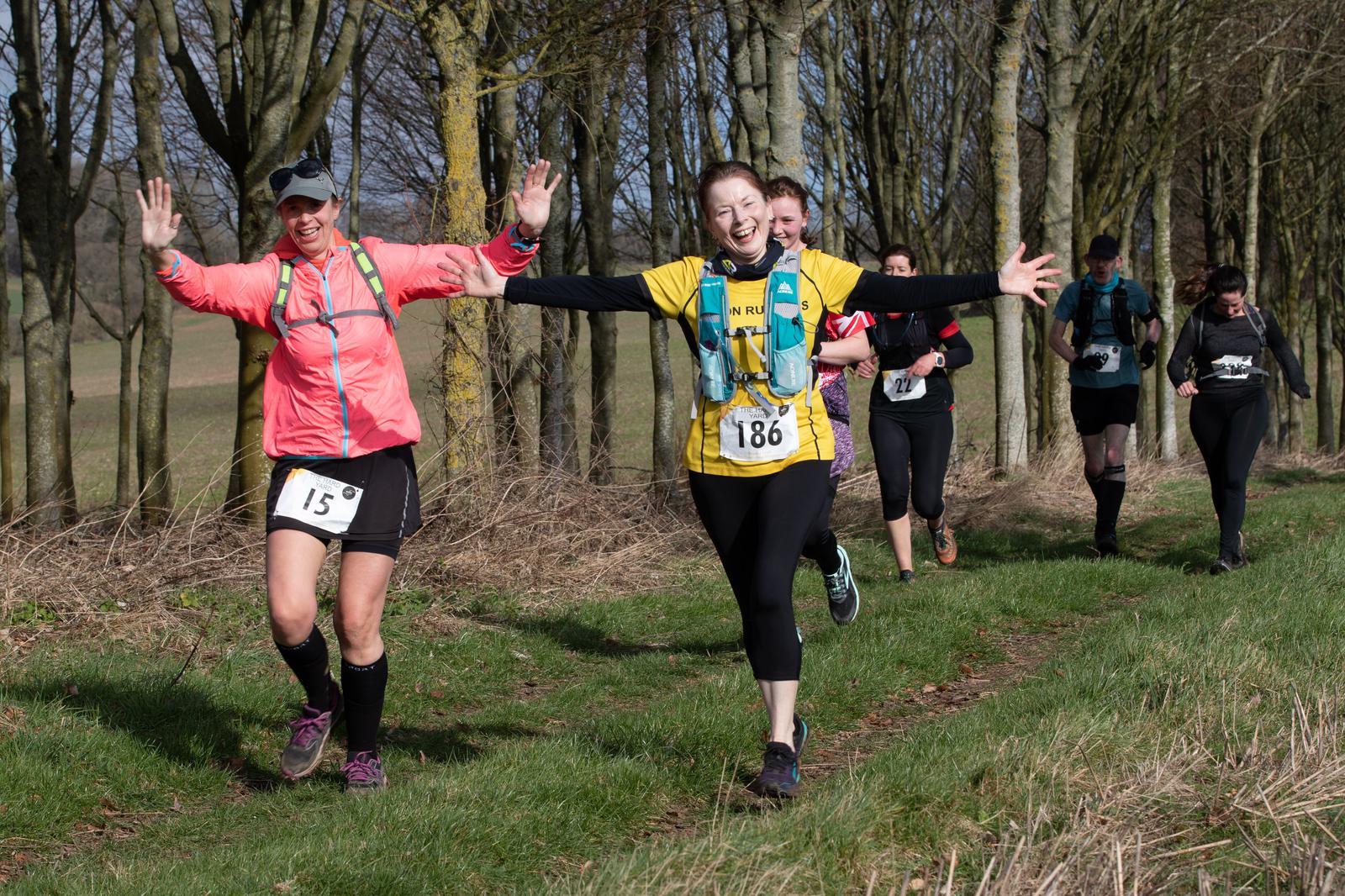 Two women in running gear, one in a pink jacket and the other in a yellow shirt, joyfully raise their arms as they run on a grassy trail. Behind them, other runners follow. Trees and shrubs line the trail, and the atmosphere is energetic and cheerful.