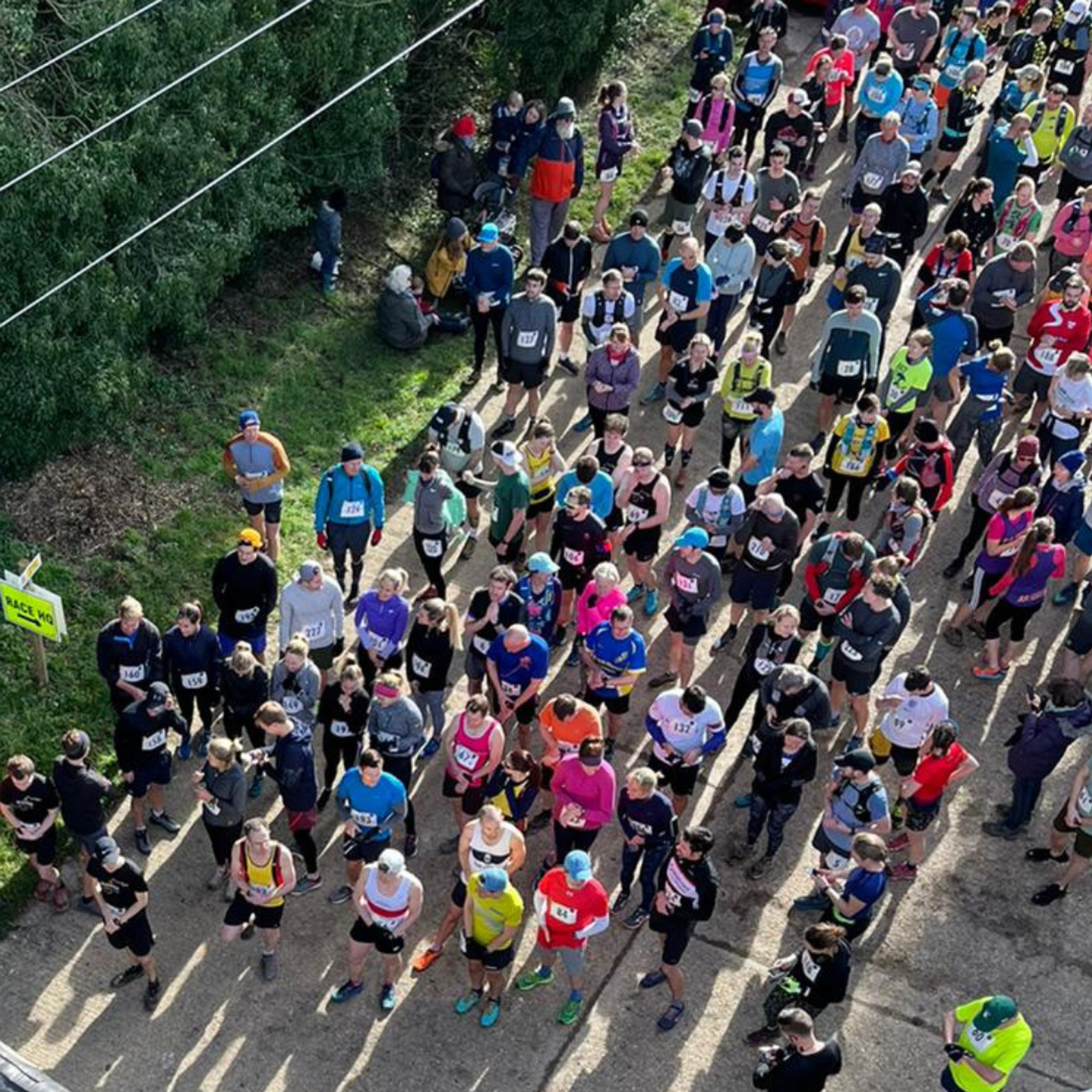 A crowd of runners gathered at the starting line of a race. The participants are dressed in athletic gear of various colors and some have race bibs pinned to their shirts. The scene is set on a paved path bordered by greenery with spectators in the background.