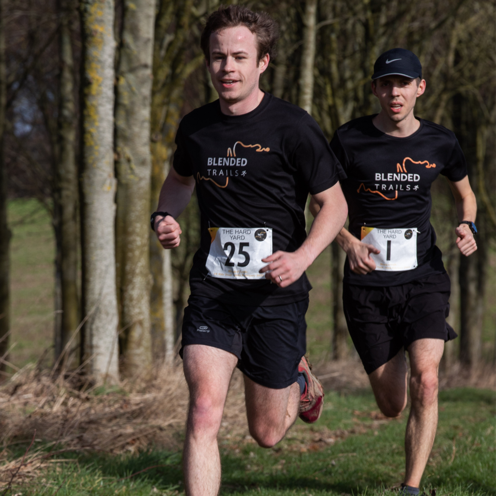 Two runners participate in a race along a wooded path, wearing black T-shirts with orange "Blended Trails" text and numbered race bibs (#25 and #1). The background features trees and grass, with sunlight filtering through. Both runners look focused.