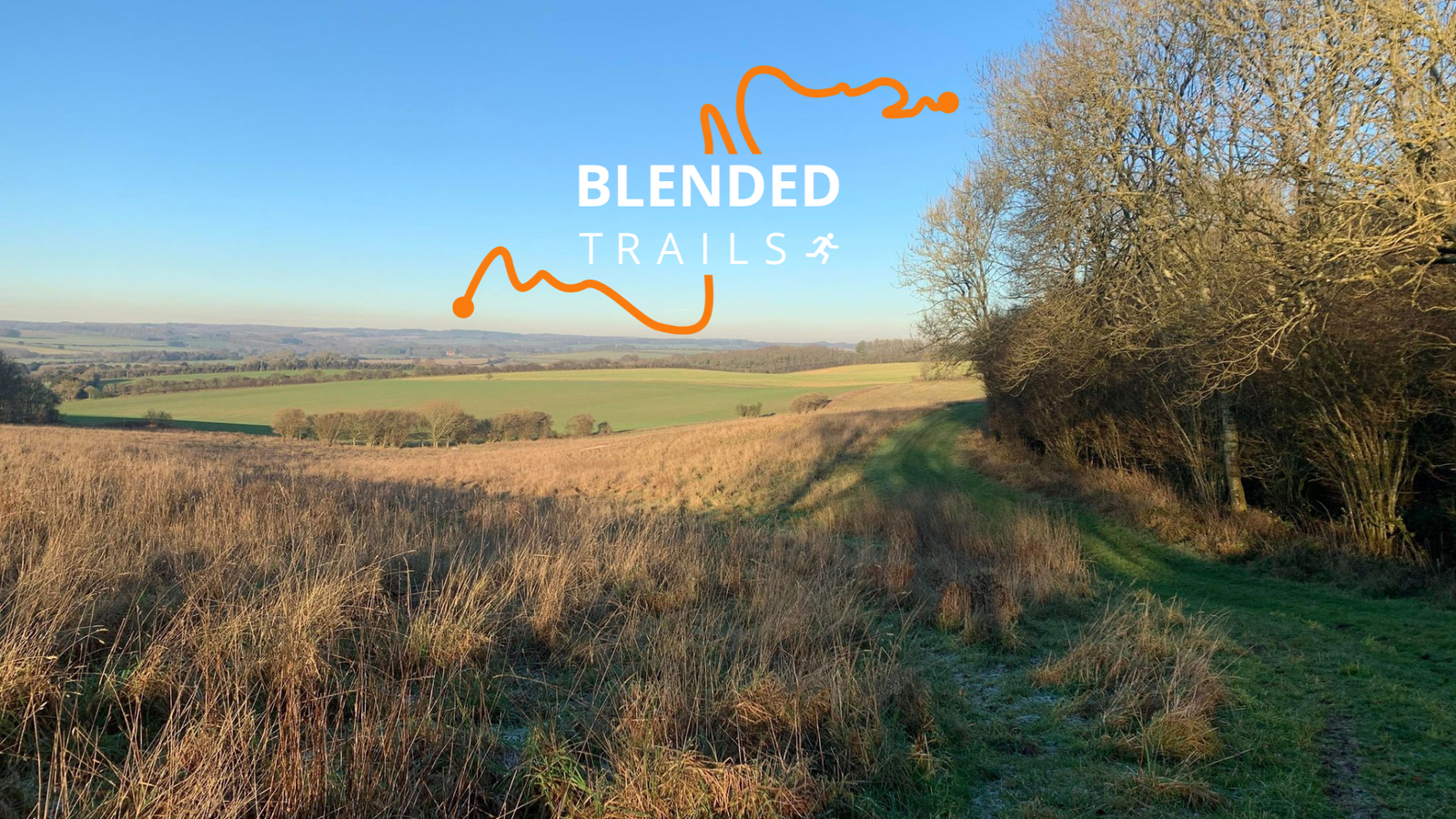 A grassy trail winds through a serene meadow and into a forested area under a clear blue sky. The text "Blended Trails" is overlaid on the image with an orange trail line and a small running figure icon. The landscape features rolling hills and distant fields.