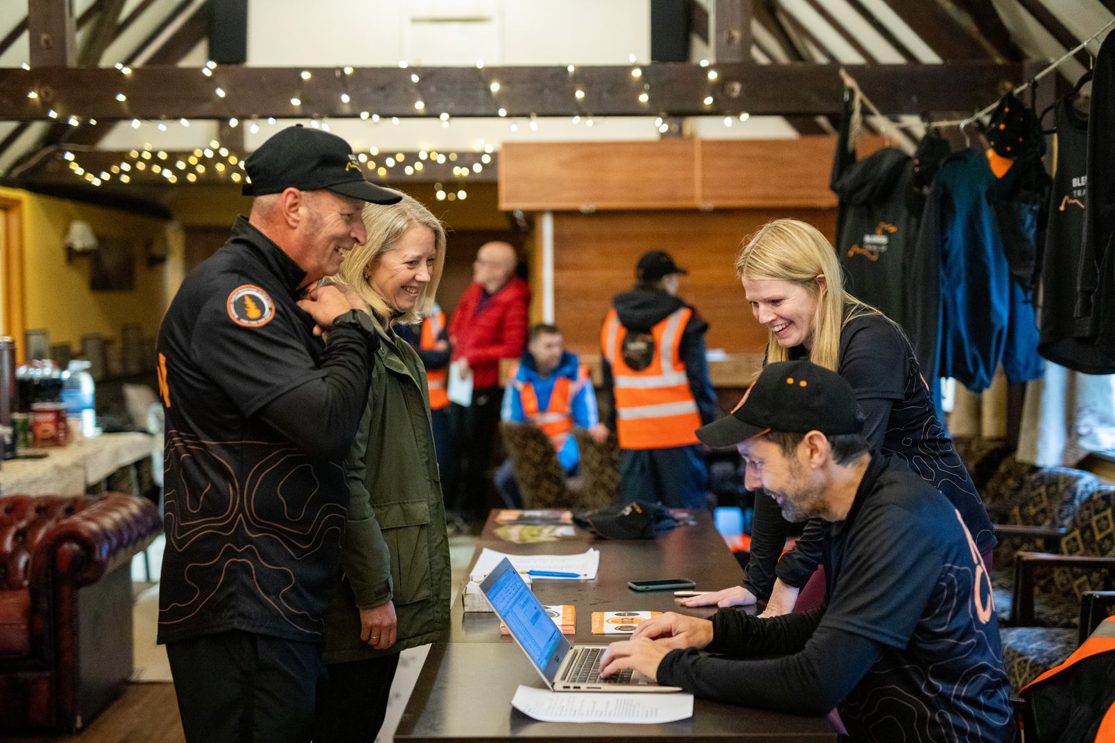 Two people smile while checking in at a registration desk manned by two staff members with a laptop, inside a cozy room decorated with string lights and jackets, with more people in orange vests in the background.