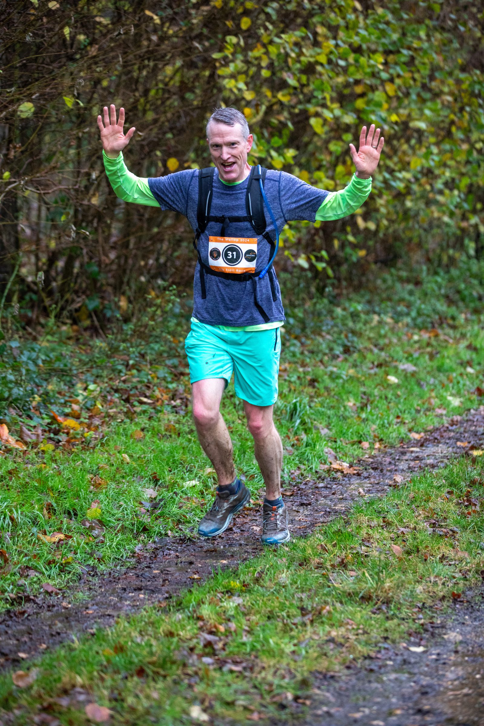 A man running on a muddy forest trail, wearing a race bib, gray t-shirt, blue shorts, and raising his hands in the air while smiling. Green foliage is visible in the background.