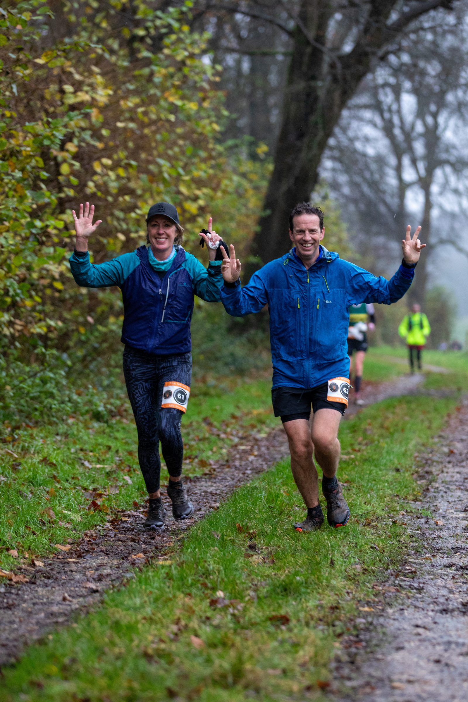 Two smiling runners in blue jackets jog on a muddy forest trail, raising their hands in celebration. Other participants are visible in the background on the overcast day.