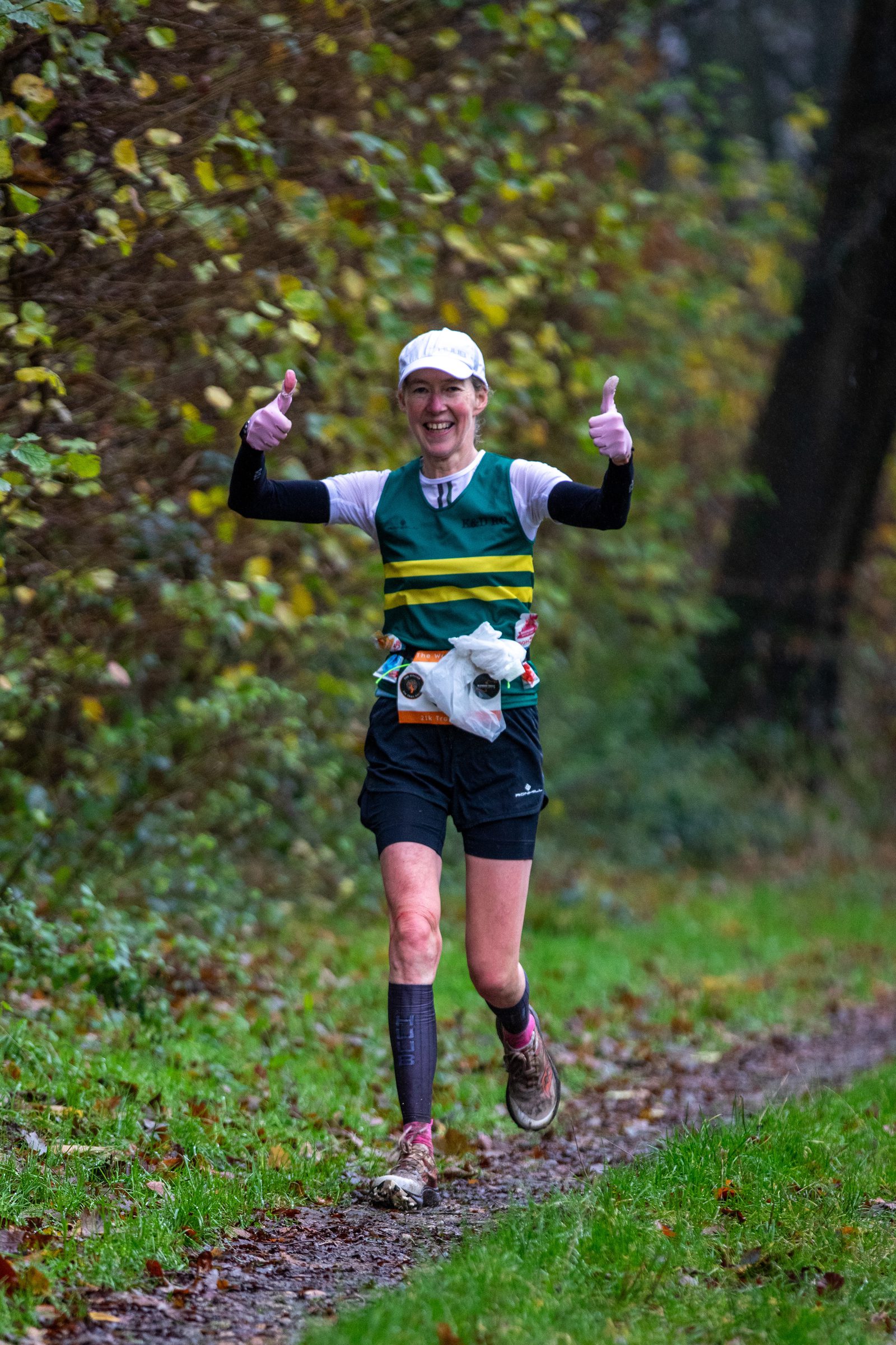 A runner smiles and gives two thumbs up while jogging on a muddy forest trail. They wear a white cap, striped green top, black shorts, and a race bib, surrounded by greenery and autumn leaves.
