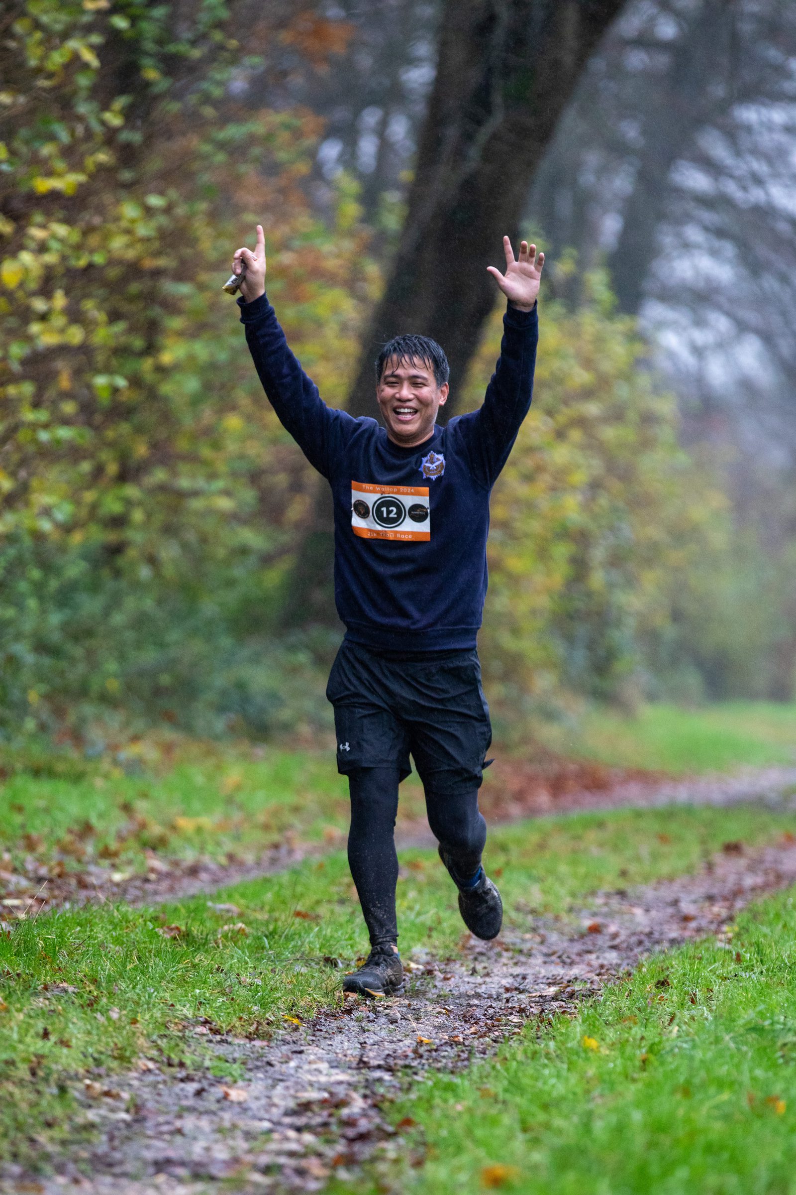 A man wearing a race bib runs on a muddy trail, raising both arms in celebration. He is smiling, surrounded by trees and greenery on a rainy day.