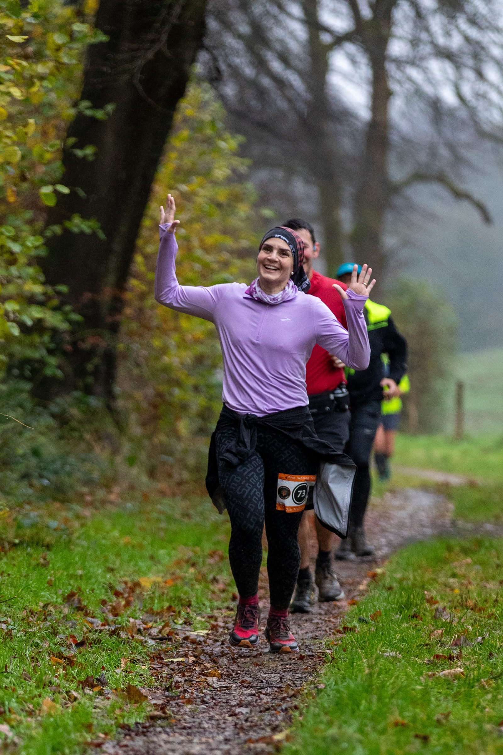 A woman in a lavender top and black leggings smiles and raises her hands while running on a forest trail, followed by other runners. The path is lined with green grass and autumn leaves.