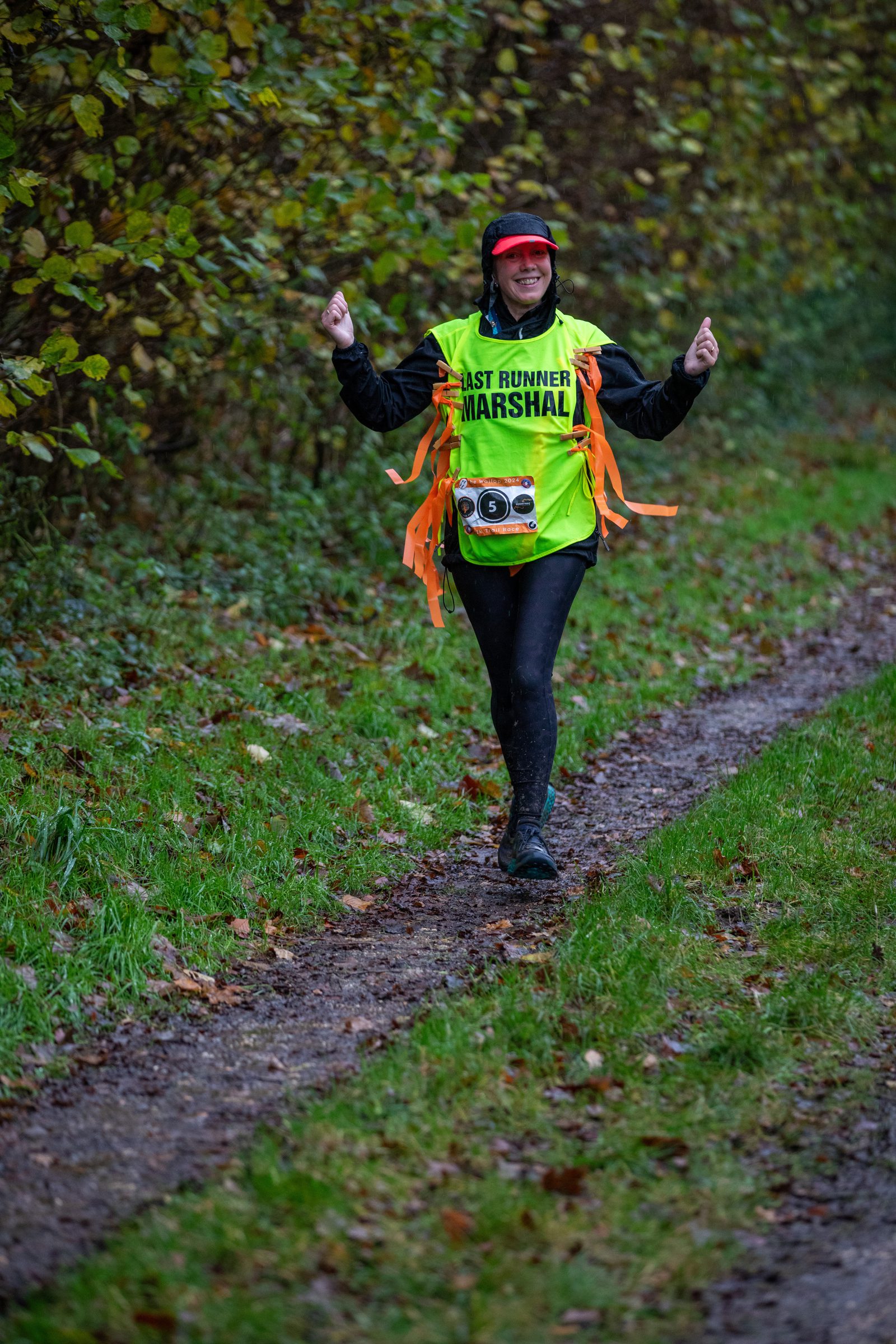 A person in a bright yellow "LAST RUNNER MARSHAL" vest walks on a muddy trail in a wooded area, smiling and giving two thumbs up, with orange ribbons and a camera around their waist.