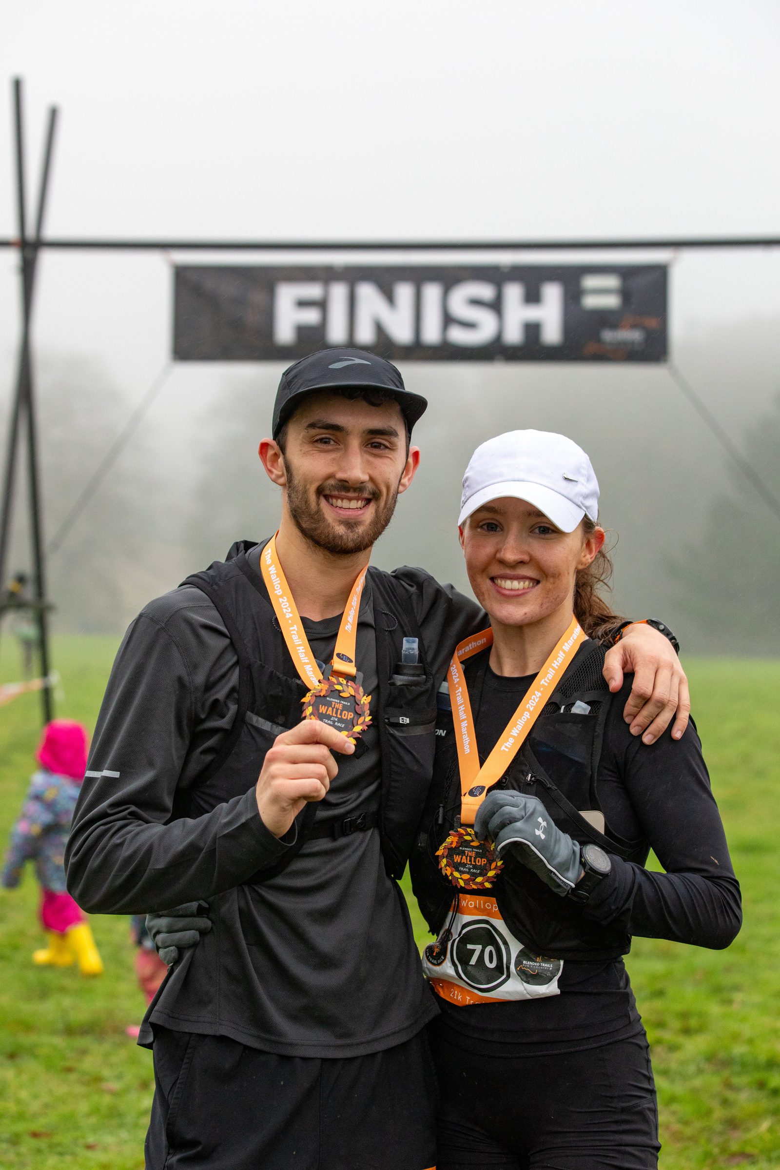 Two smiling runners wearing medals stand together in front of a "Finish" banner on a foggy day. Both are dressed in dark athletic gear, and one wears a white cap. A few people are visible in the background.