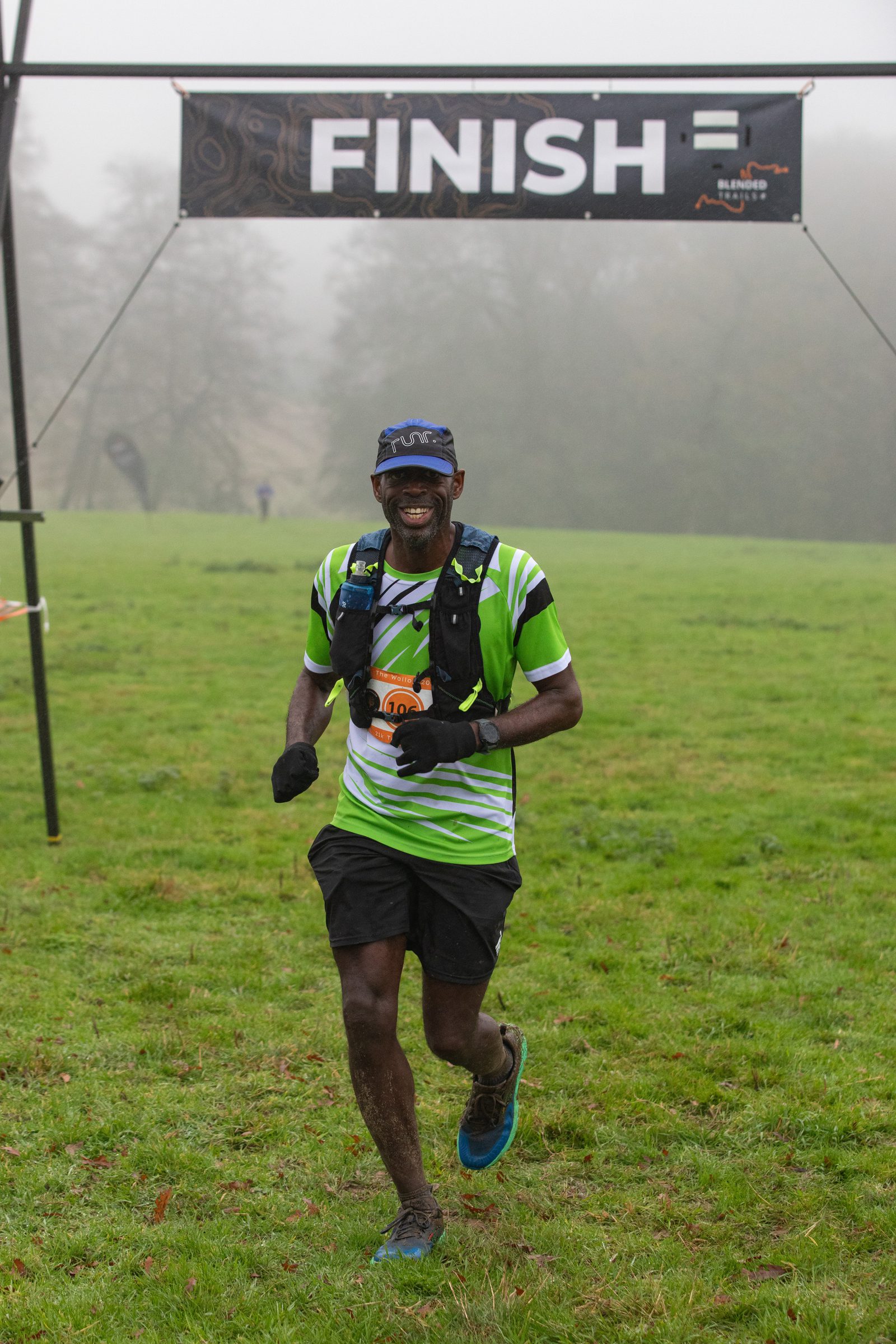 A smiling runner in a green and white striped shirt, black shorts, and a cap crosses a grassy finish line, with a "FINISH" banner overhead on a foggy day.