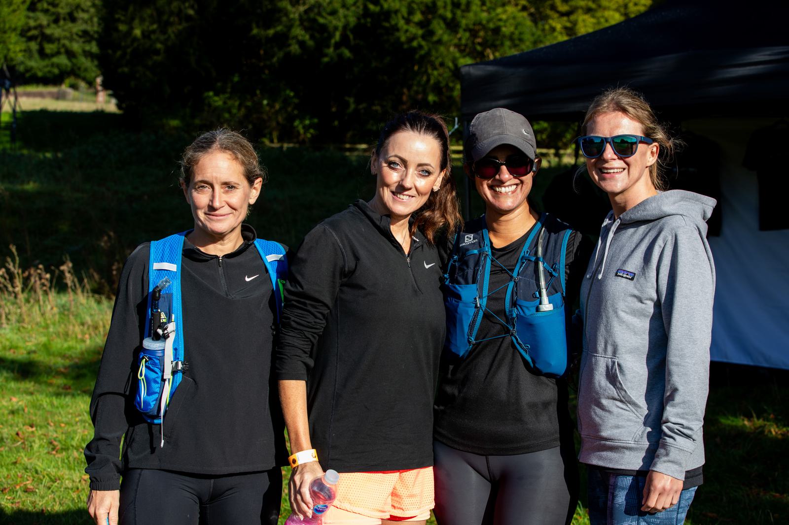 Four women, dressed in casual athletic wear, stand side by side outdoors on a sunny day. Two are wearing hydration packs and one holds a water bottle, while the other two wear sunglasses. They're all smiling, with trees and a black canopy visible in the background.