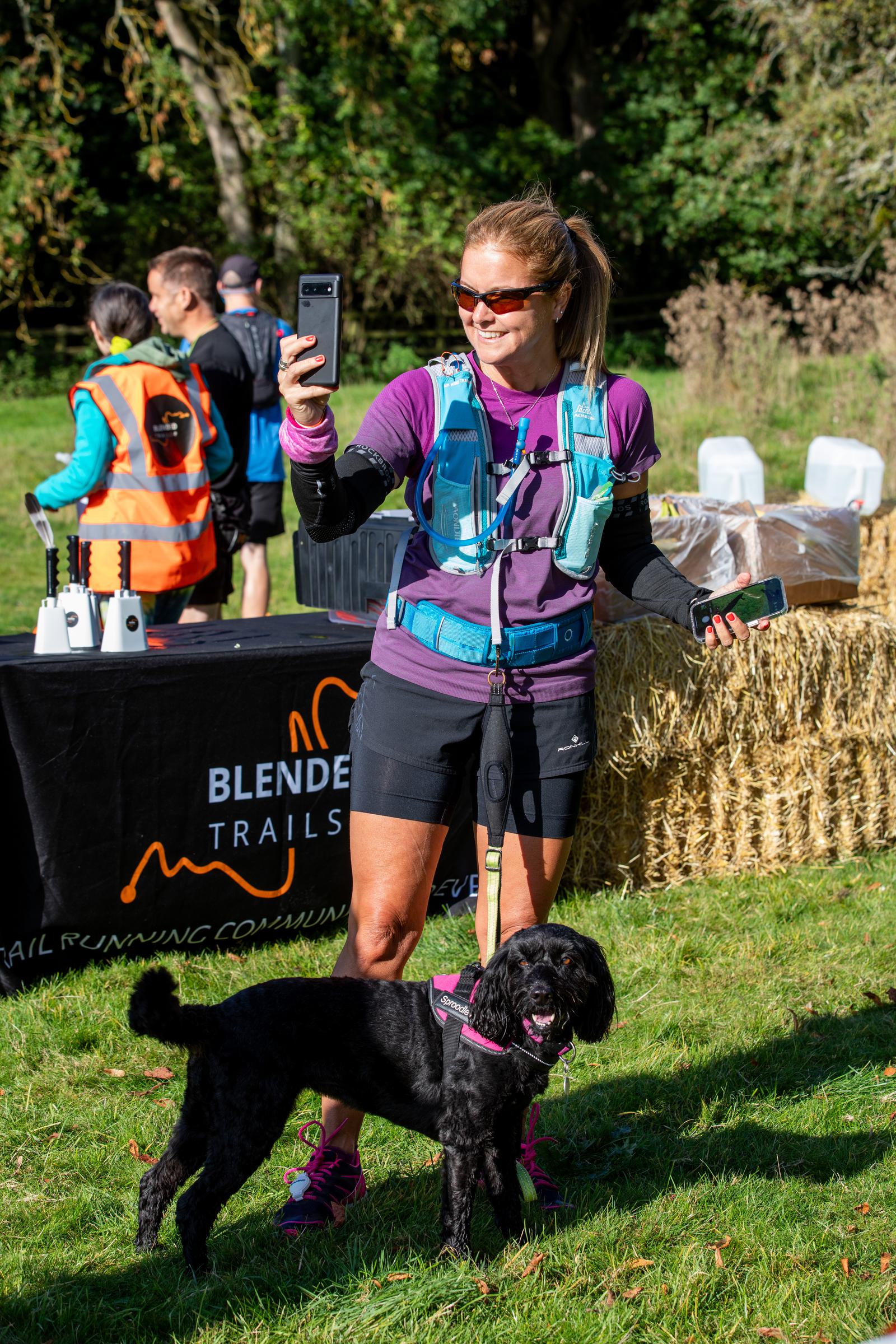 A woman in athletic gear, wearing a hydration pack and sunglasses, takes a selfie while holding a phone in the other hand. Beside her stands a black dog on a leash. Behind them, blurry figures and a table covered with equipment are visible.