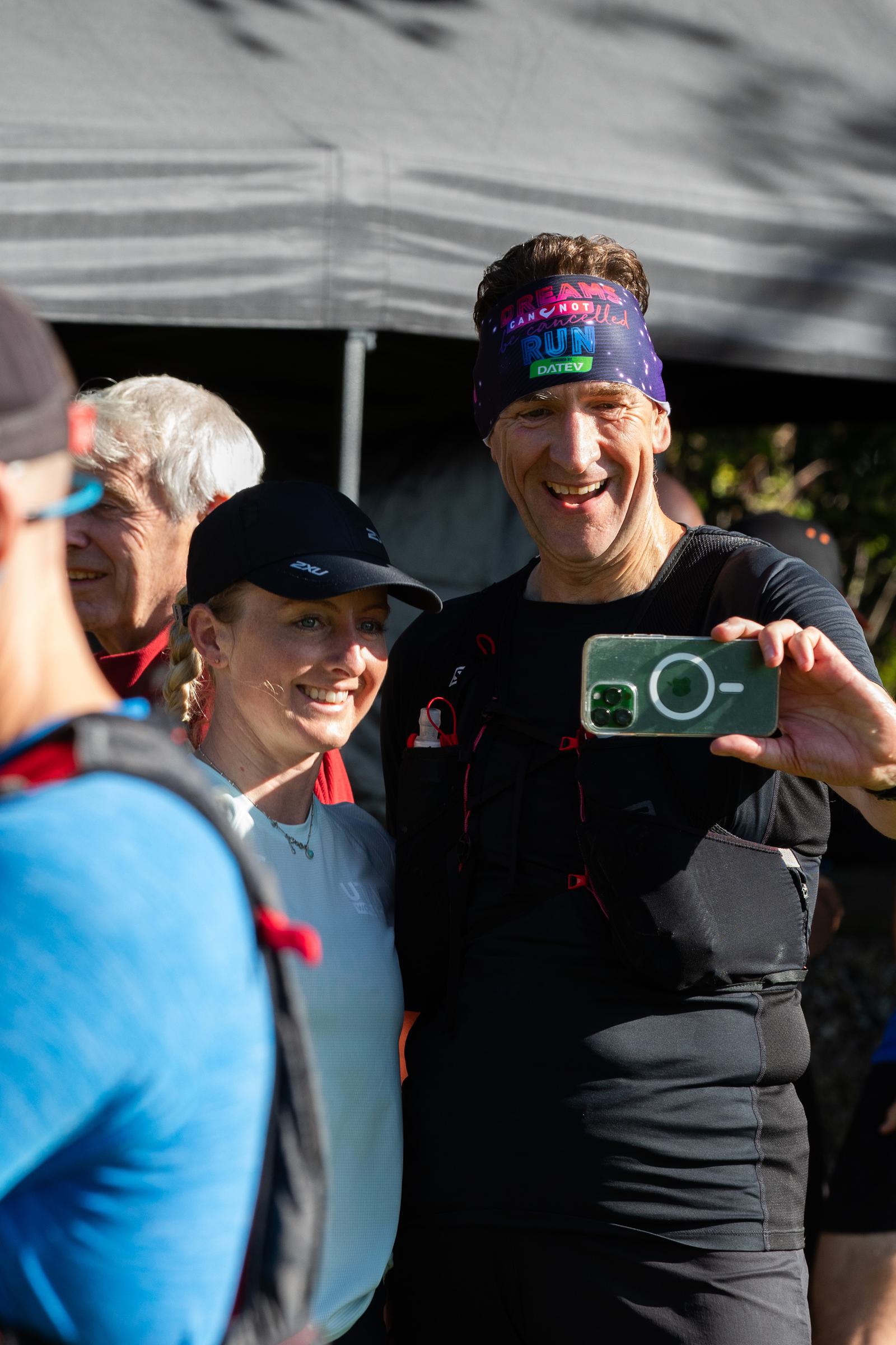 A man and woman in athletic gear take a selfie together outdoors. The man is holding up a green smartphone, and he is wearing a headband with colorful text. Other people are partially visible in the background. They appear to be at a sports event or gathering.