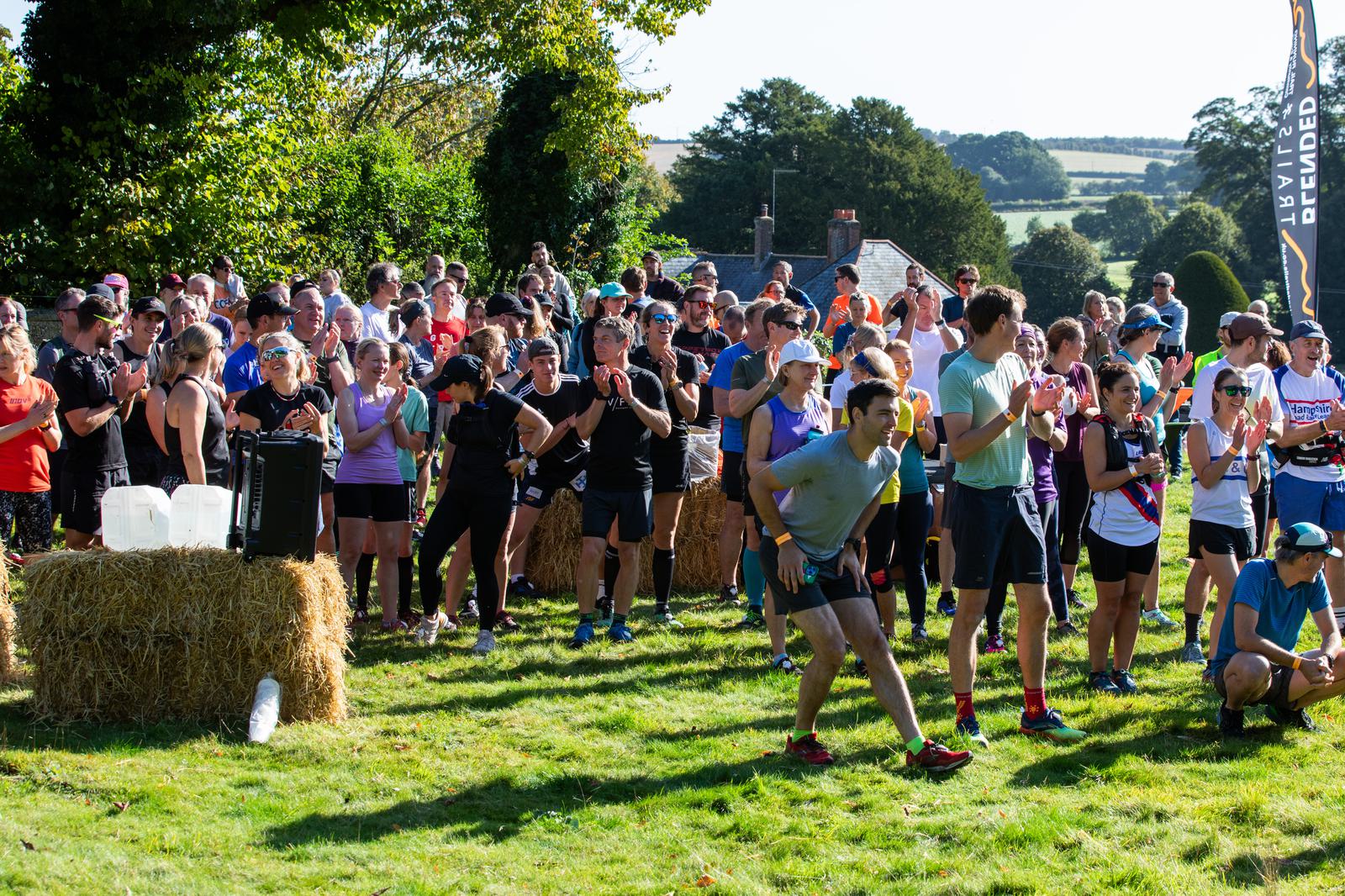 A large group of runners gathered in an outdoor grassy area, some clapping and cheering. Hay bales and a table with water stations are visible. Trees and a scenic landscape can be seen in the background. The weather appears sunny and pleasant.