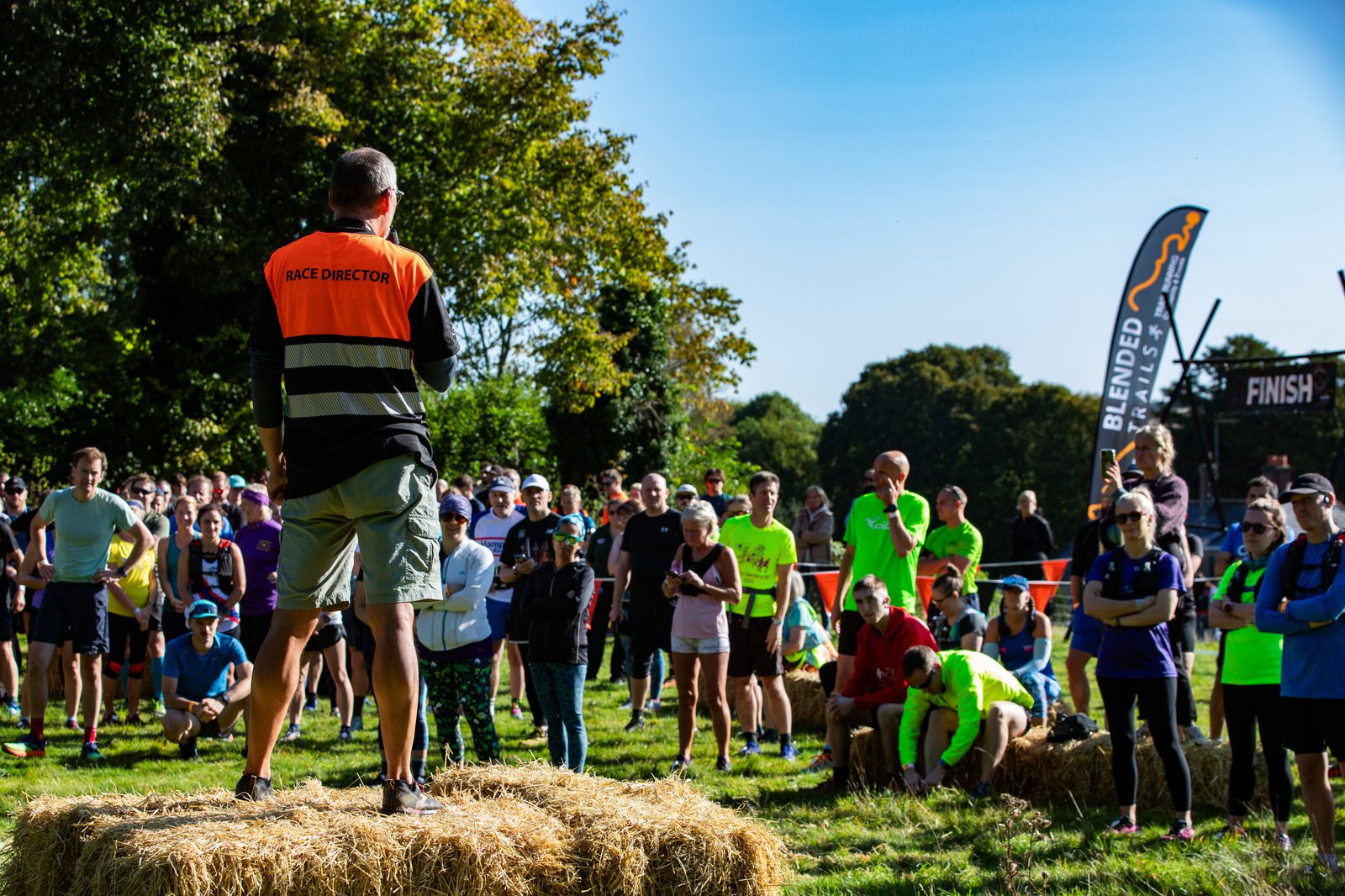 A race director in an orange vest addresses a crowd of runners gathered on a grassy field. Several participants are sitting on hay bales, listening attentively. It's a sunny day with green trees in the background, and a "Finish" sign is visible.