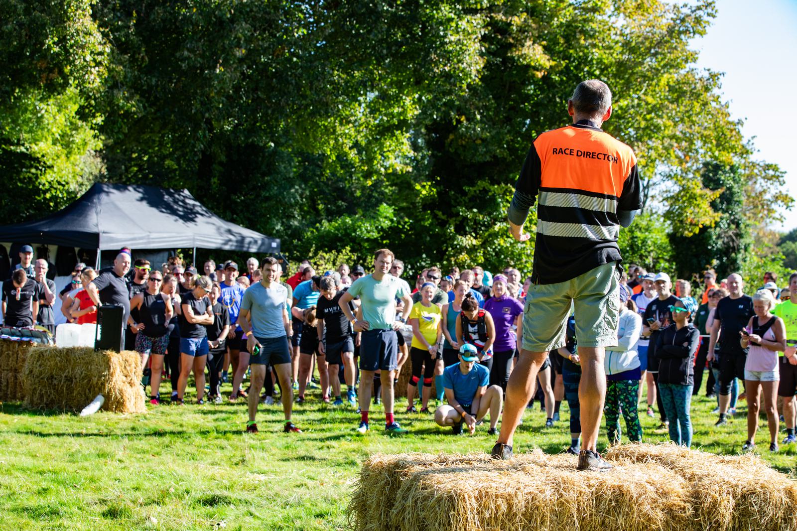 A race director wearing an orange vest stands on a hay bale addressing a crowd of participants in a park. The group listens attentively, with some people in running attire. Trees and a canopy tent are visible in the background, indicating an outdoor event.