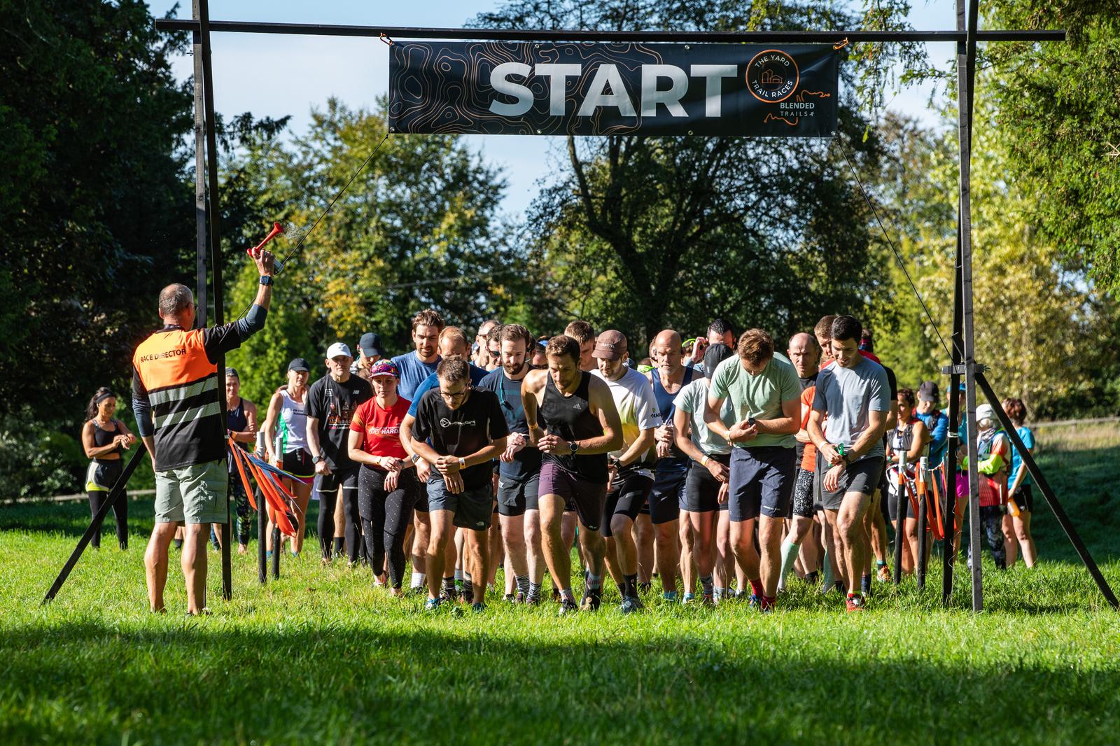 A group of runners gathers at the start line of a race in a park, with a person in an orange vest signaling the beginning using a whistle. The start sign hangs overhead, and the background shows lush green trees and a sunny sky.
