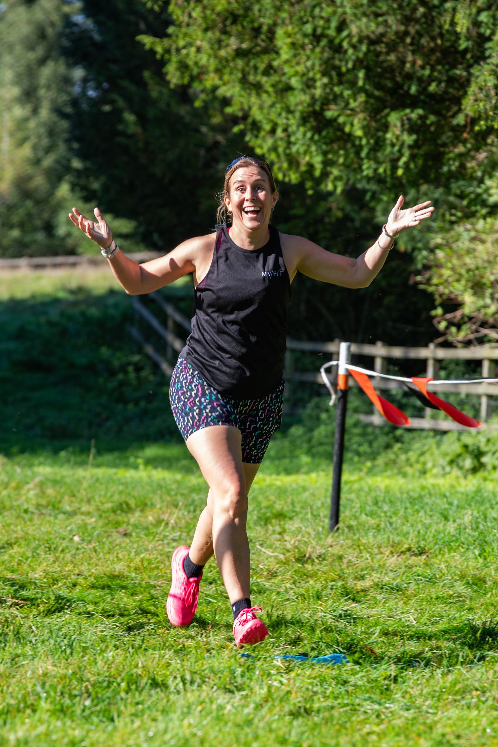 A woman wearing a black tank top and patterned shorts runs on a grassy path with her arms raised and a joyful expression. She wears pink running shoes and a headband. Trees and a fence are visible in the background. The sun is shining brightly.