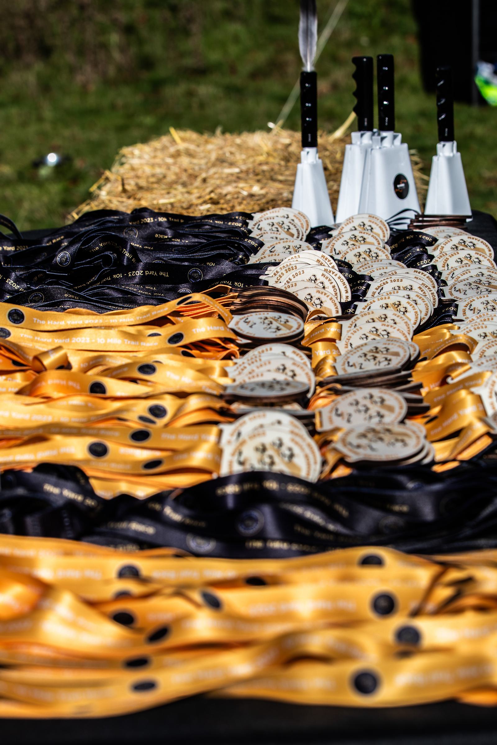 A table is filled with numerous gold and black ribbons attached to medals, arranged in rows. Behind the medals, white cowbells with black handles are stacked on straw bales. The background features green grass, hinting at an outdoor event.