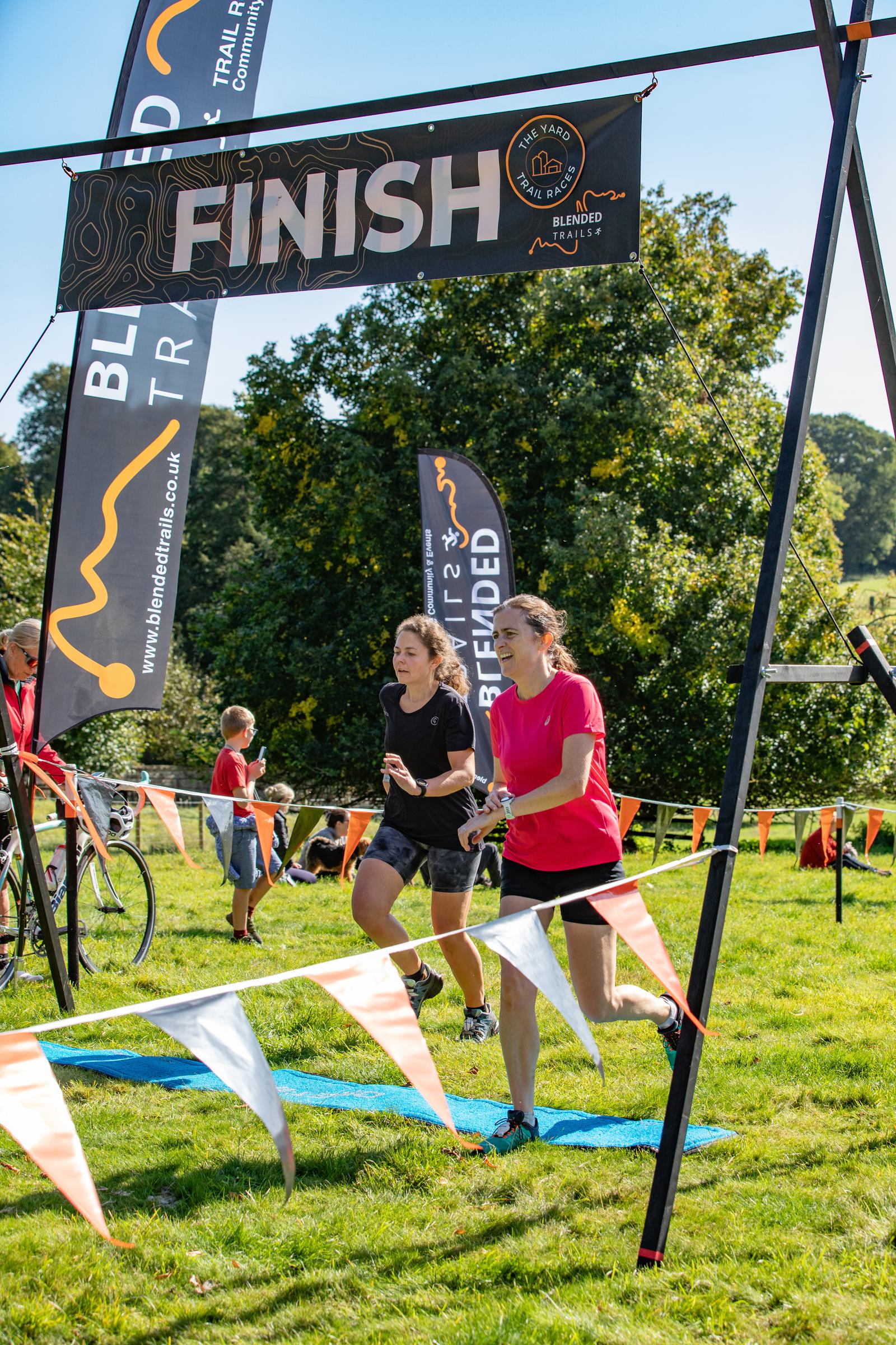 Two women are crossing the finish line of a race. They are running on grass under a banner that reads "FINISH," with event flags and trees in the background. Both are smiling and appear to be in motion. The weather is sunny and clear.