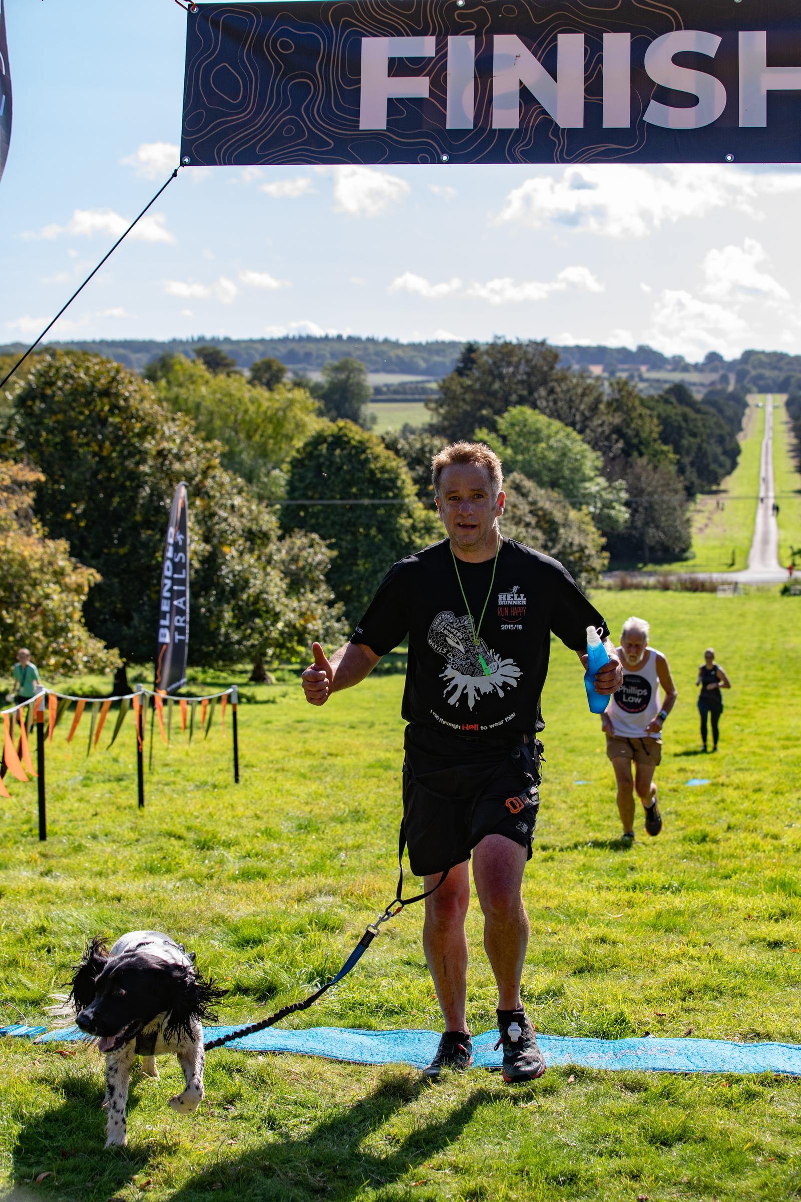 Runner crosses a grassy finish line with a dog on a leash, both appearing joyful. A "FINISH" banner is overhead. Another runner is seen in the background under a sunny sky, with a tree-lined path visible in the distance.