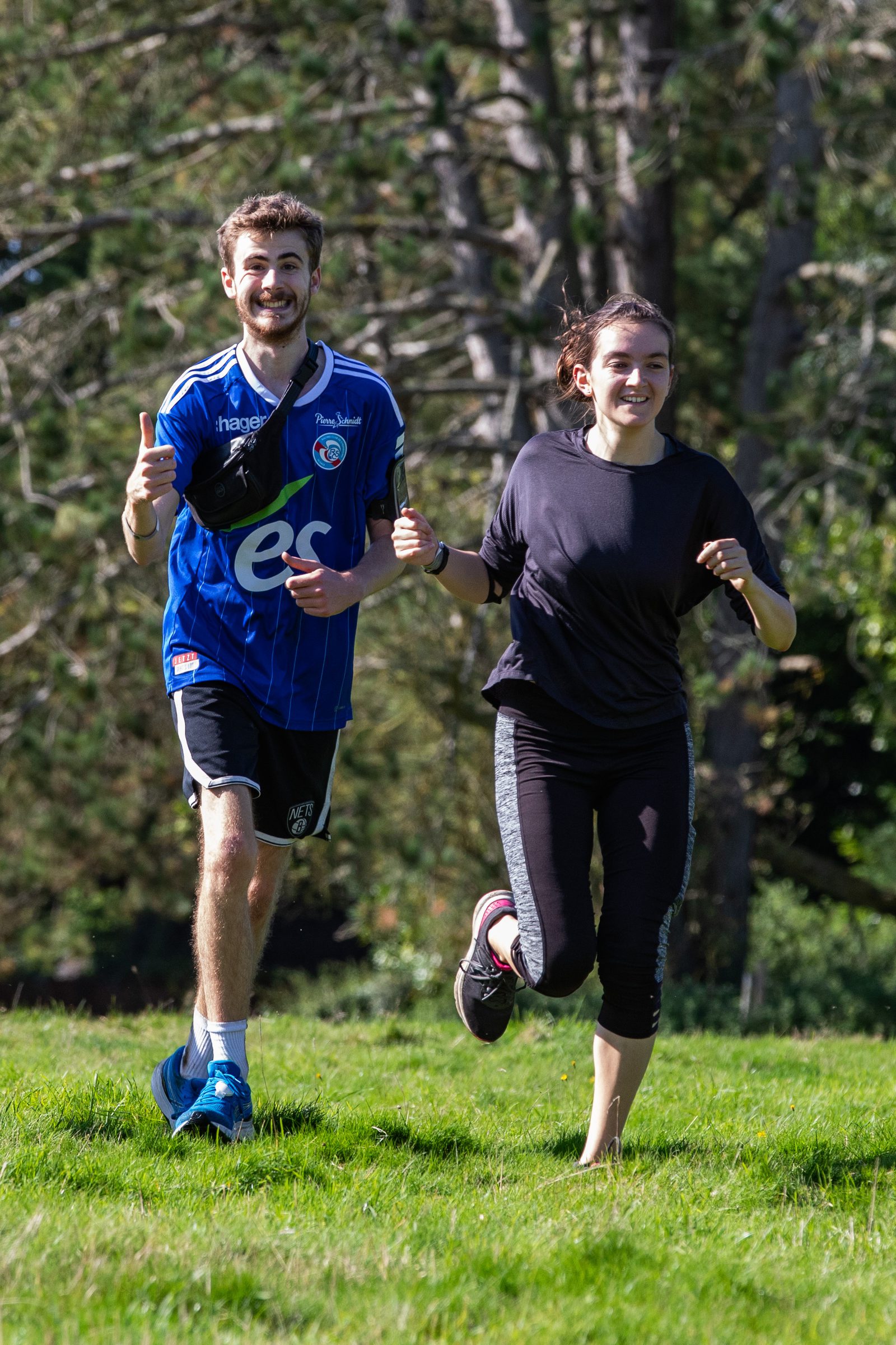 Two people are running on a grassy field while smiling; the person on the left is giving a thumbs-up and wearing a blue sports jersey, black shorts, and blue shoes. The person on the right is wearing a black top and pants. Trees and foliage are visible in the background.