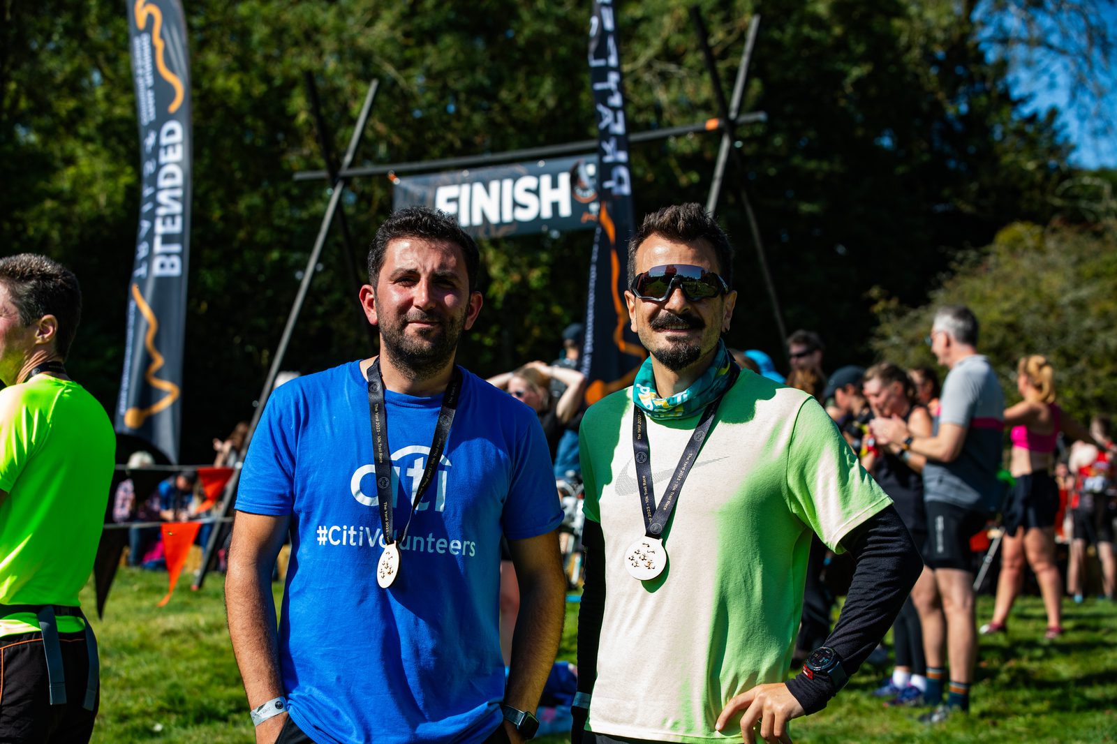Two men stand proudly with medals around their necks at a race finish line, smiling at the camera. The man on the left wears a blue shirt, while the man on the right wears a light green shirt and sunglasses. Other runners and spectators are seen in the background.