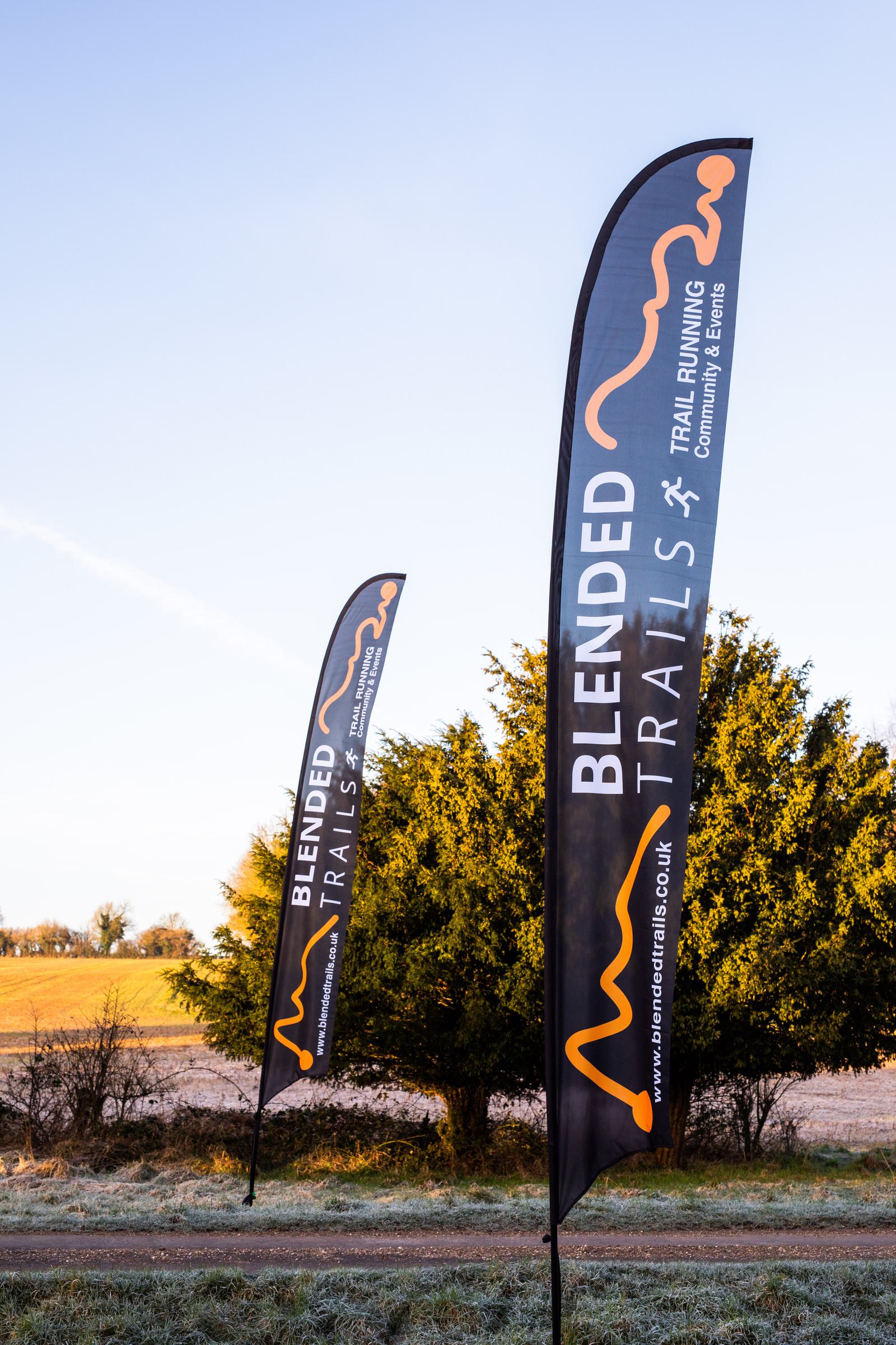 Two black vertical flags with orange and white text reading "Blended Trails" and "Trail Running Events" are standing in a grassy area with bushes and trees in the background. The sky is clear, and the sun is illuminating the flags and landscape.
